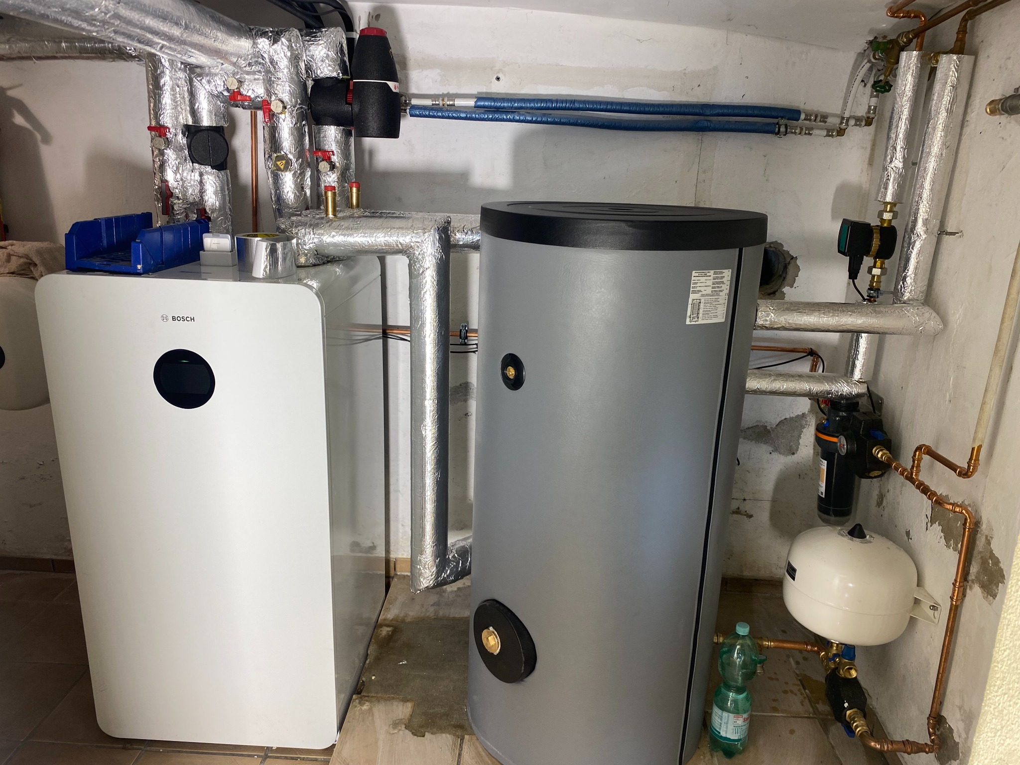 Kotłownia z białym kotłem Bosch, szarym zasobnikiem ciepłej wody, rurami w izolacji aluminiowej i instalacją hydrauliczną z miedzianych rur.