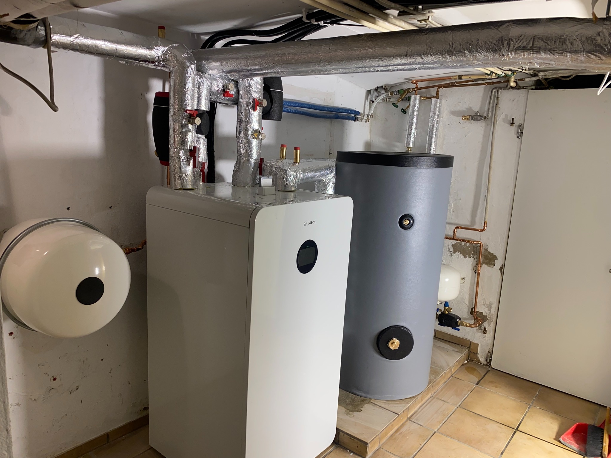 Biała pompa ciepła Bosch z szarym zbiornikiem akumulacyjnym w piwnicy, rury w izolacji aluminiowej, płytki podłogowe w odcieniach beżu.