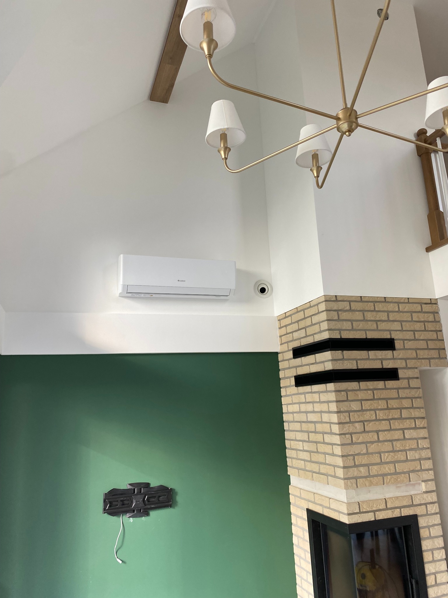 Wnętrze domu z zamontowaną klimatyzacją typu split, widoczny również kominek z jasnej cegły, złoty żyrandol i uchwyt na telewizor na zielonej ścianie.