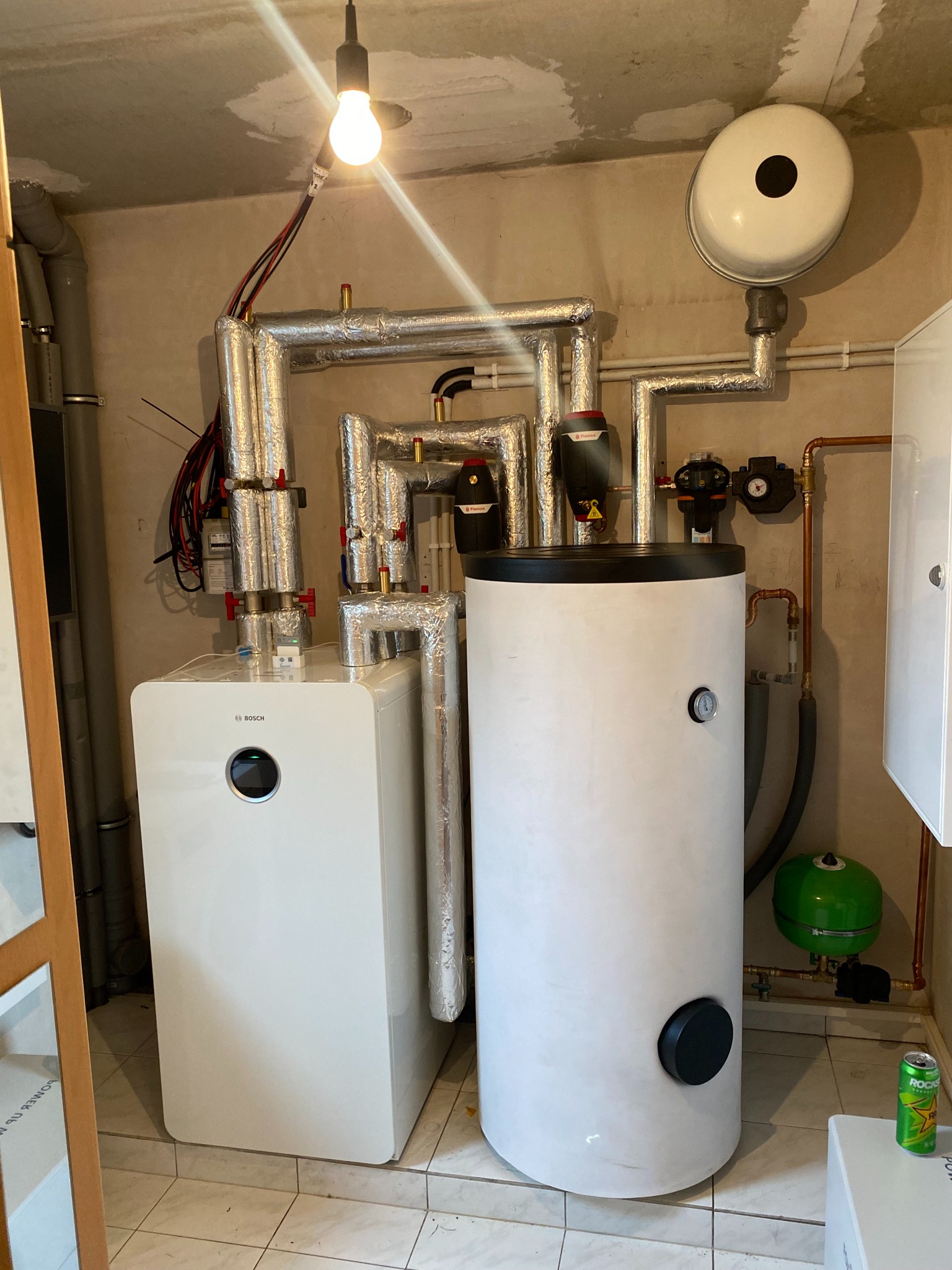 Biały, nowoczesny zestaw grzewczy Bosch z zbiornikiem akumulacyjnym, rury w izolacji aluminiowej, mały zielony zbiornik wyrównawczy, widoczna puszka napoju energetycznego obok zbiornika.