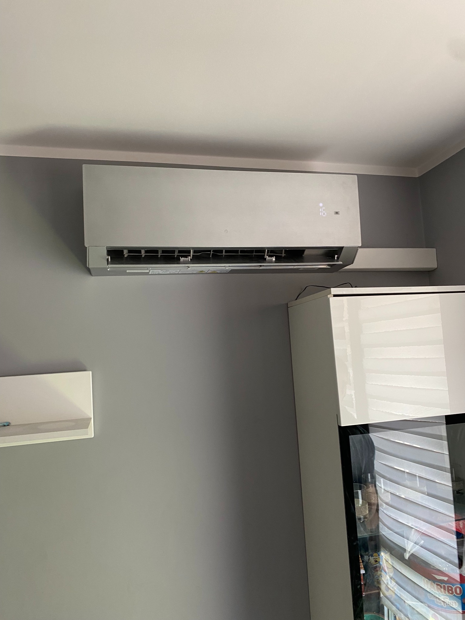 Szary klimatyzator ścienny z wyświetlaczem temperatury 16 stopni, zamontowany w szarym pokoju nad białą szafką.