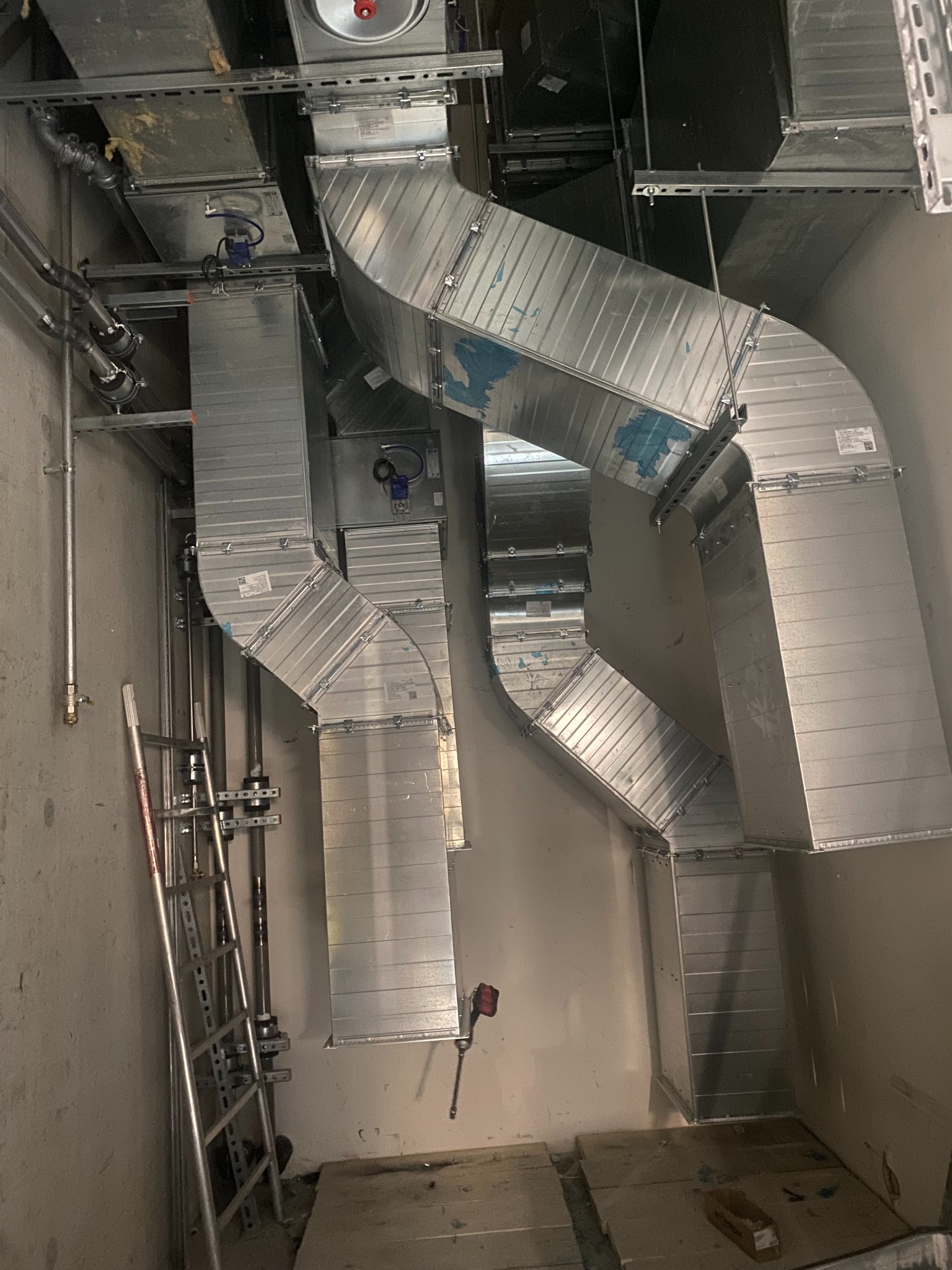 Widok z dołu na system kanałów wentylacyjnych z blachy ocynkowanej, zamontowany pod sufitem w surowym wnętrzu, z widoczną drabiną i rurami instalacyjnymi.