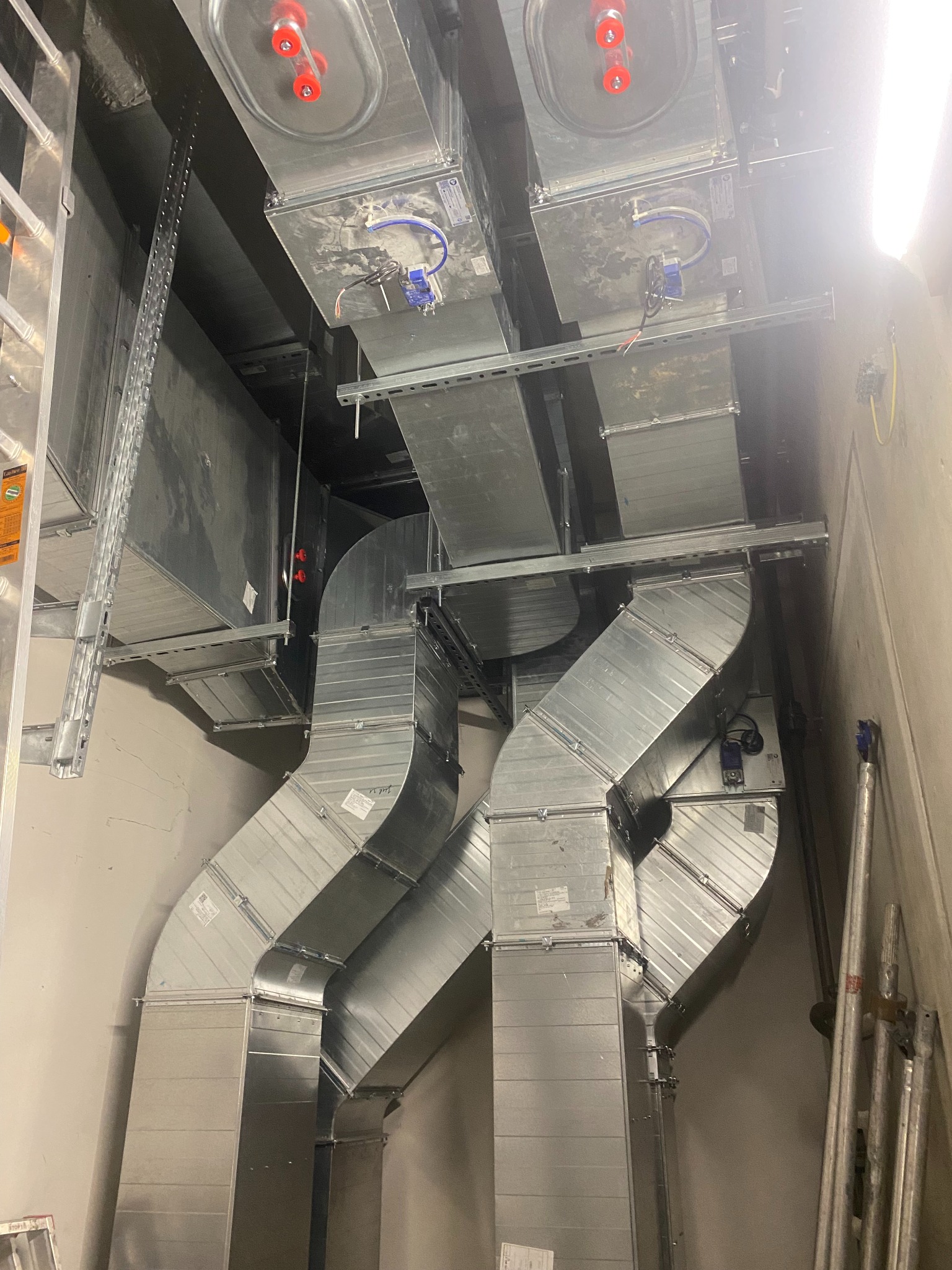 Metalowe kanały wentylacyjne zamontowane pod sufitem, widoczne elementy mocujące i przewody elektryczne, drabina oparta o ścianę.