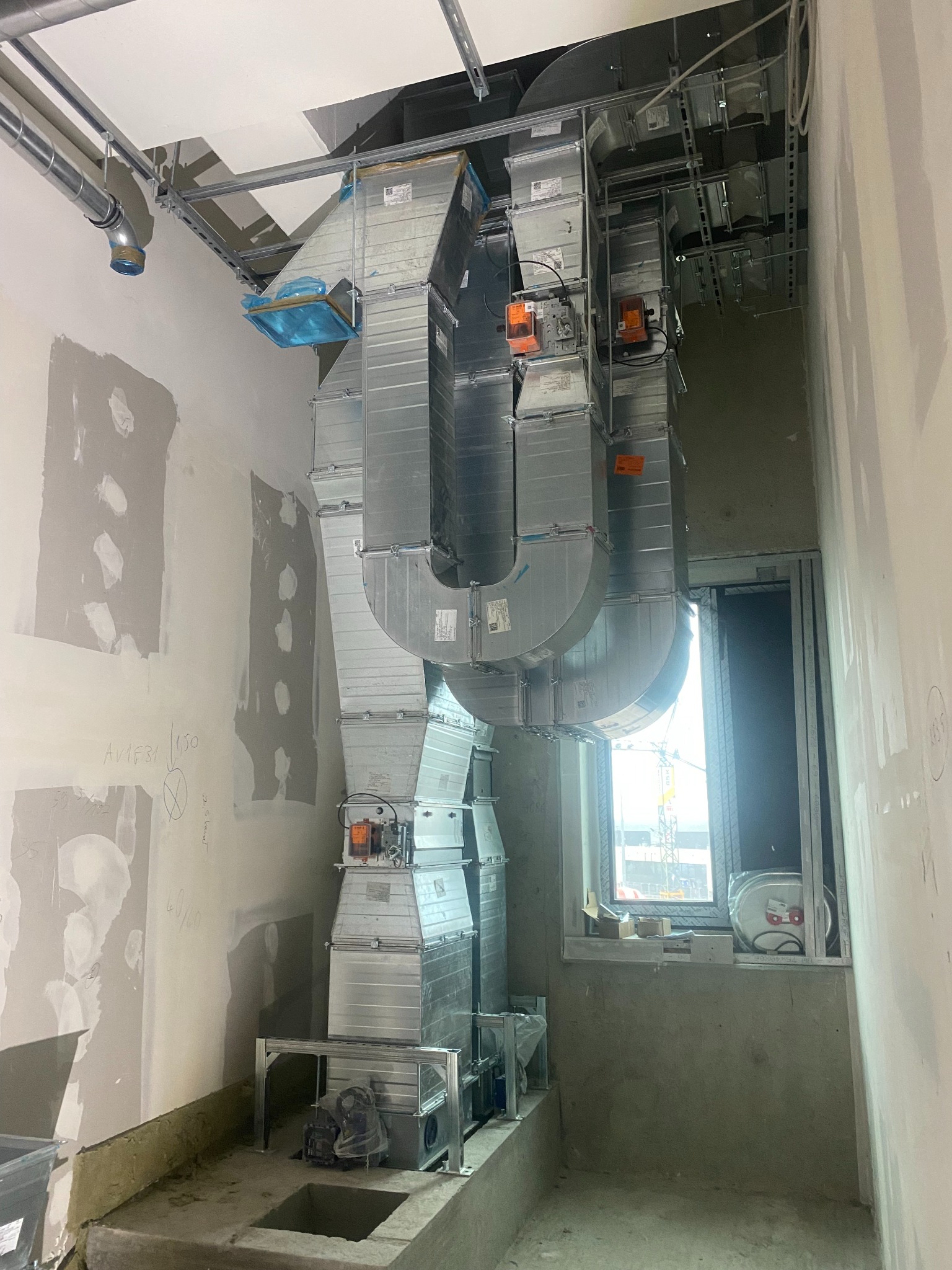 Instalacja systemu wentylacji z metalowych kanałów w surowym wnętrzu budynku w trakcie budowy, widoczne elementy montażowe i okno z widokiem na plac budowy z dźwigiem.
