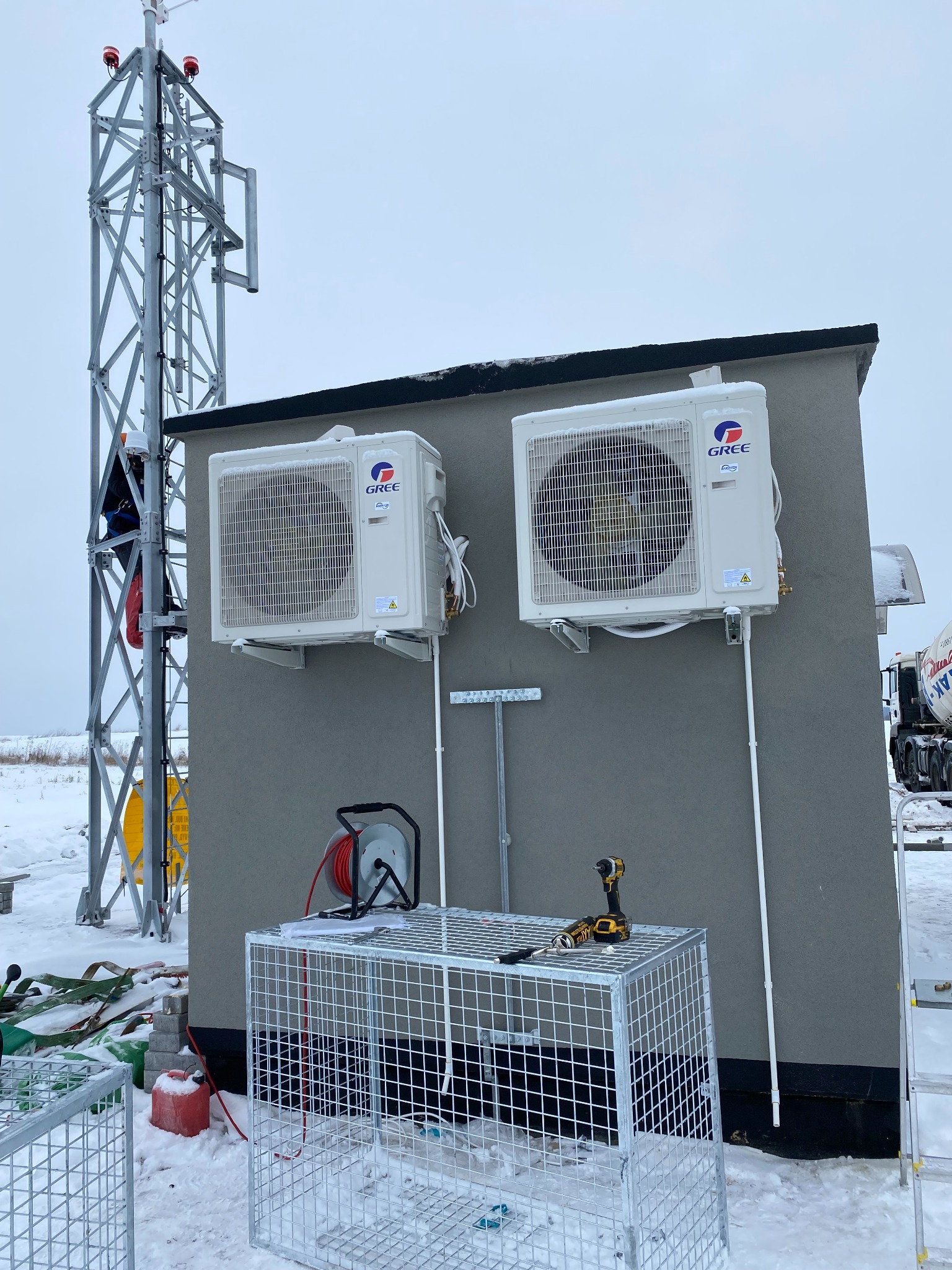 Dwa klimatyzatory marki Gree zamontowane na szarej ścianie budynku, widoczne narzędzia na metalowej klatce, śnieg na dachu i ziemi, w tle wieża telekomunikacyjna z pracownikiem na wysokości...