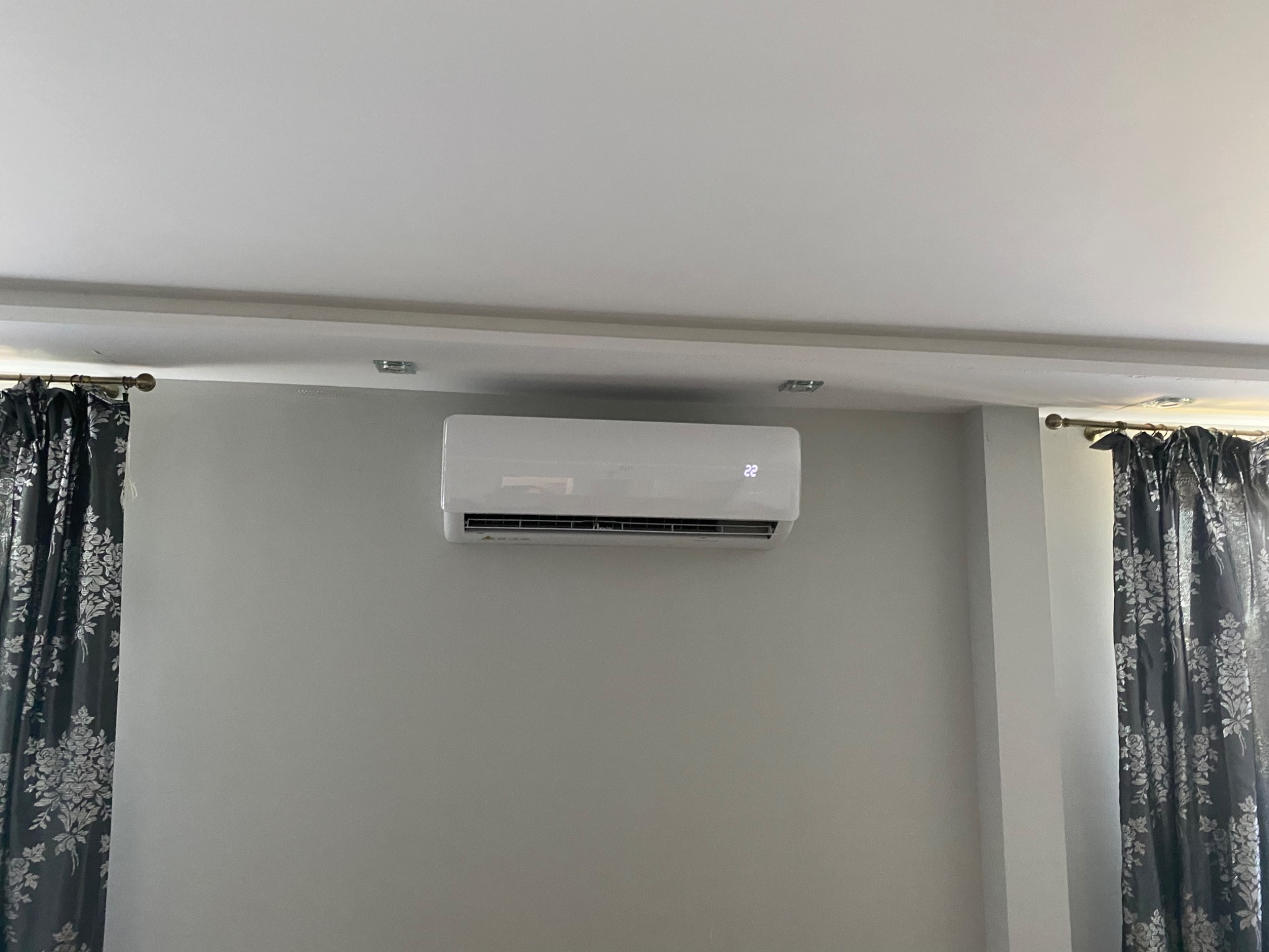 Biała klimatyzacja ścienna z wyświetlaczem temperatury 22 stopnie, zamontowana na szarej ścianie w pokoju z zasłonami w kwiaty.