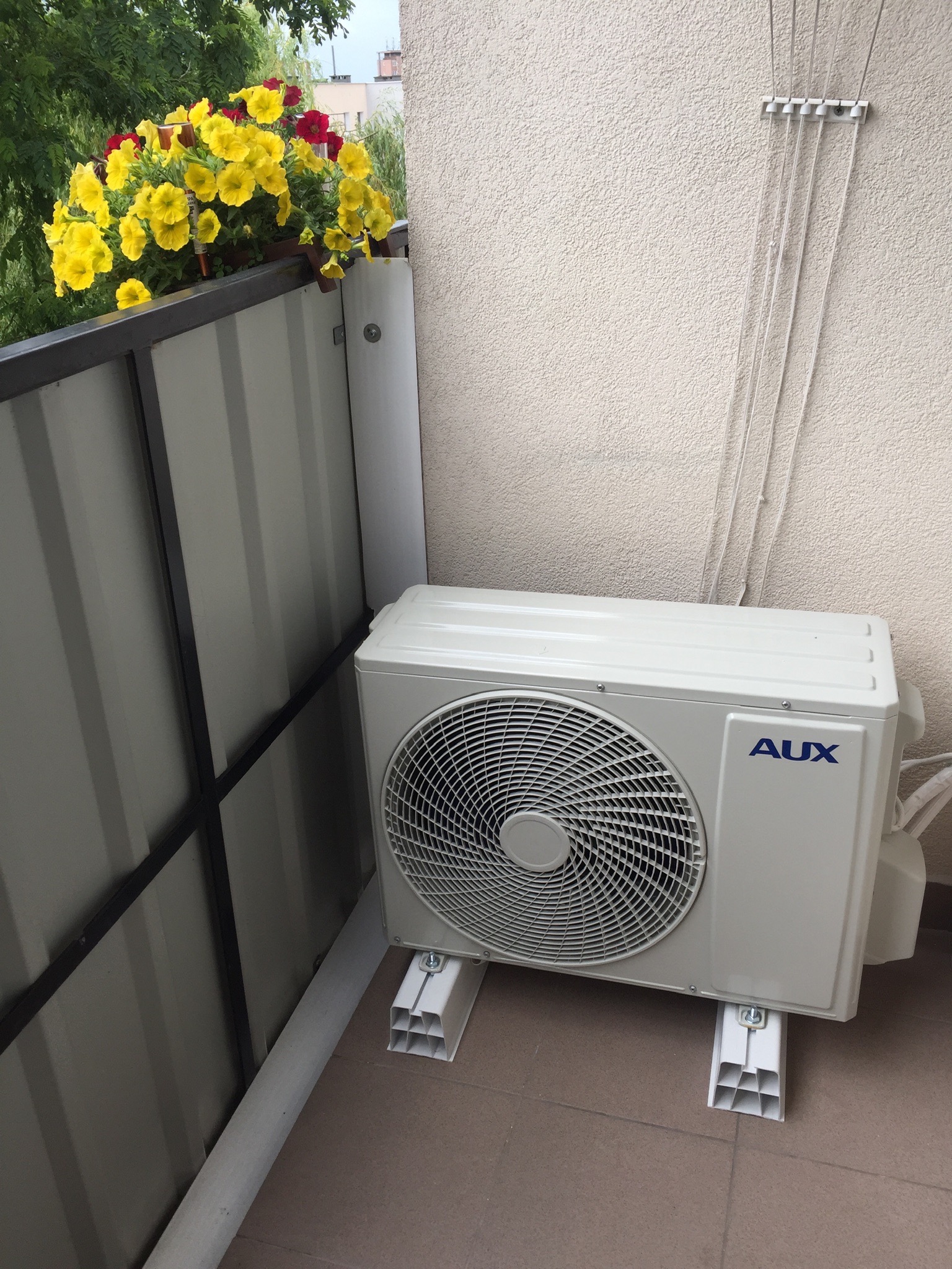 Zewnętrzna jednostka klimatyzacji marki AUX zamontowana na balkonie, widok z boku, z kwiatami w tle.