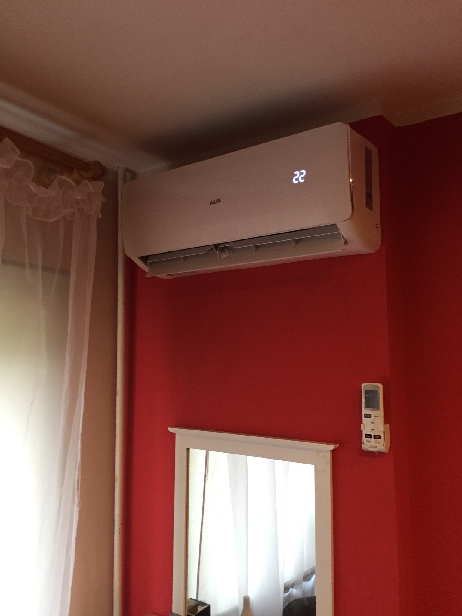 Biały klimatyzator AUX zamontowany na czerwonej ścianie w pokoju, obok okna z firanką i lustra w białej ramie, z pilotem przytwierdzonym do ściany.