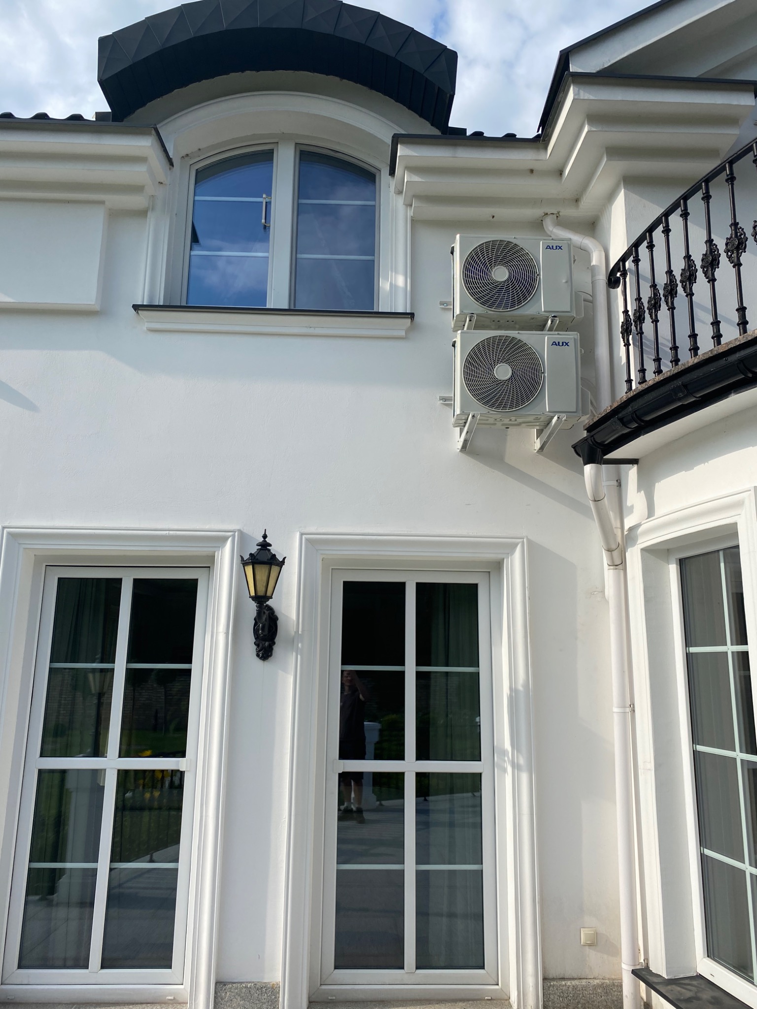 Dwa białe klimatyzatory marki AUX zamontowane na jasnej elewacji domu z oknami w białych ramach i czarnym, ozdobnym oświetleniem zewnętrznym.