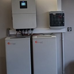 EKOLOGICZNY DOM - Dwa białe magazyny energii Felicitysolar z falownikiem na ścianie w pomieszczeniu. Widoczna skrzynka elektryczna i oświetlenie. Na dole adres strony internetowej.