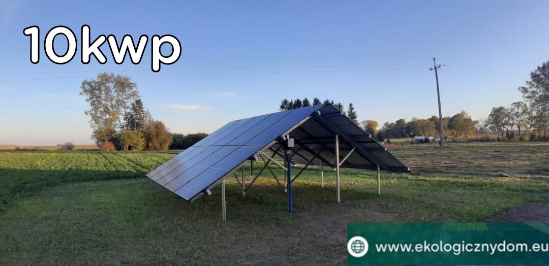 Instalacja fotowoltaiczna o mocy 10 kWp na metalowej konstrukcji ustawiona na zielonym polu z widocznymi drzewami w tle, błękitne niebo.