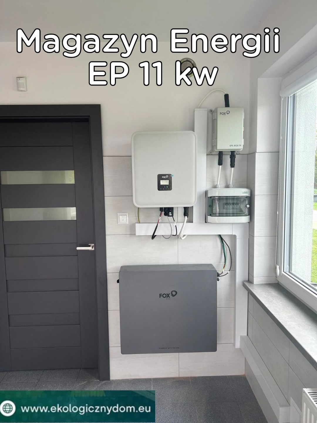 Domowa instalacja magazynu energii EP 11 kW marki FOX, zamontowana na ścianie z jasnymi płytkami, widoczna obok ciemnych drzwi i okna.