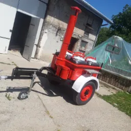 Czerwony, mobilny piec polowy na przyczepie, po renowacji, z białymi błotnikami i kominem, na tle budynku gospodarczego i foliowego tunelu.