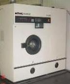 Profesjonalna pralnicza maszyna czyszcząca odzież firmową, widoczny okrągły otwór wsadowy i panel sterowania z wyświetlaczem, biała obudowa w surowym pomieszczeniu.