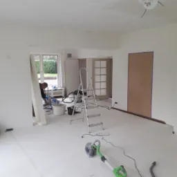 Przygotowanie do malowania: szlifierka do gładzi na podłodze, drabina, wiadro z farbą i malarz w tle w jasnym, przestronnym wnętrzu z zabezpieczoną folią podłogą i oknem.