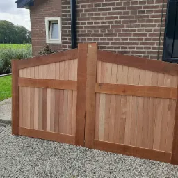 Nowe, dwuskrzydłowe, drewniane wrota ogrodowe z pionowych desek, o brązowych ramach, leżące na żwirowym podłożu przed ceglanym budynkiem z widocznym oknem i zielonym polem w tle.