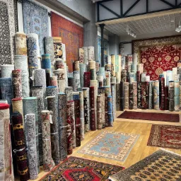 Wnętrze sklepu z dywanami: stosy zwiniętych wykładzin i dywanów z orientalnymi wzorami, ułożone na drewnianej podłodze, z dywanami rozwiniętymi na podłodze, oświetlenie punktowe.