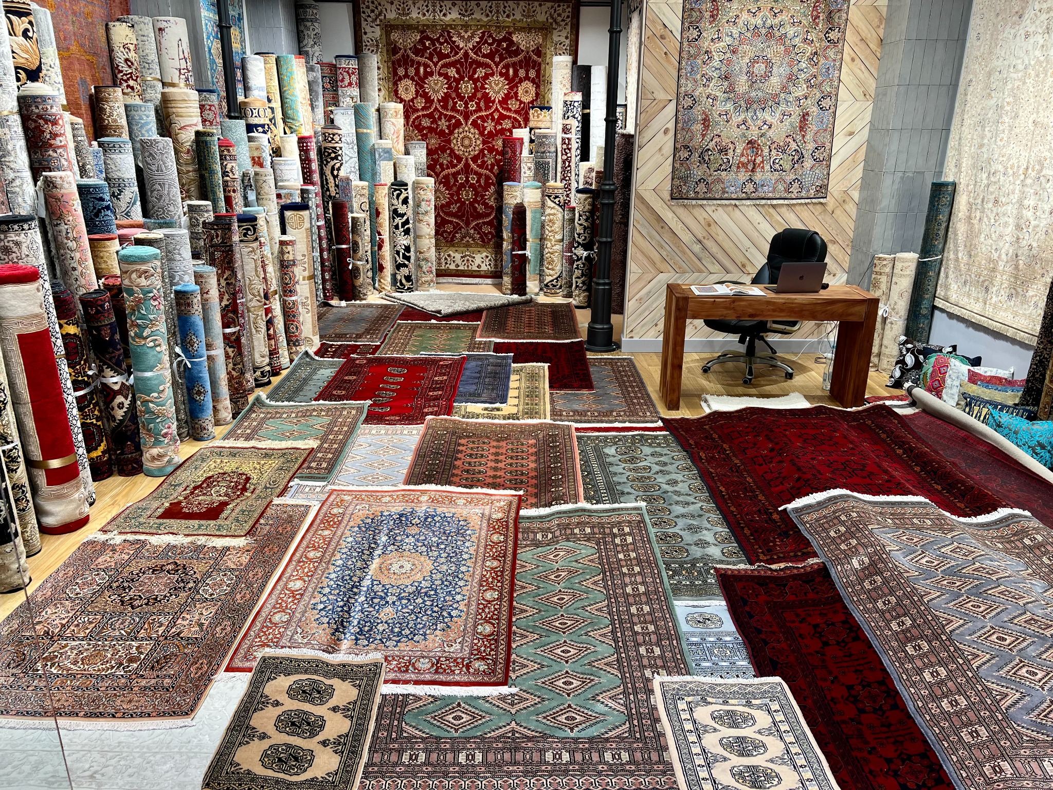 Sklep z dywanami w Warszawie: bogaty wybór wzorów i kolorów, dywany rozwinięte na podłodze i zwinięte w rulony ustawione jeden obok drugiego, widoczny fragment biurka z laptopem w tle.