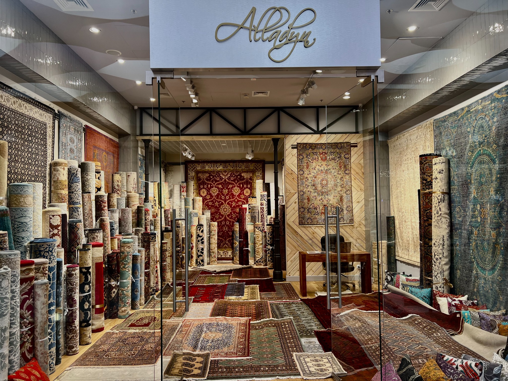 Wnętrze sklepu z dywanami: bogaty wybór zwiniętych i rozłożonych dywanów orientalnych, widoczne logo sklepu nad wejściem.