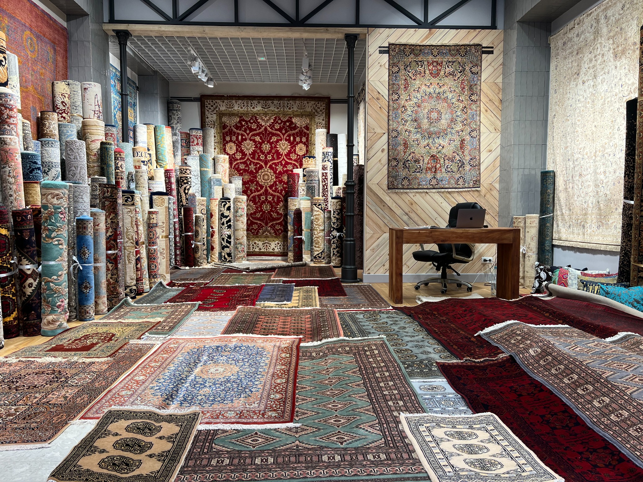 Salon z dywanami: zwinięte dywany ułożone w stosy, rozłożone dywany na podłodze, biurko z laptopem w tle.