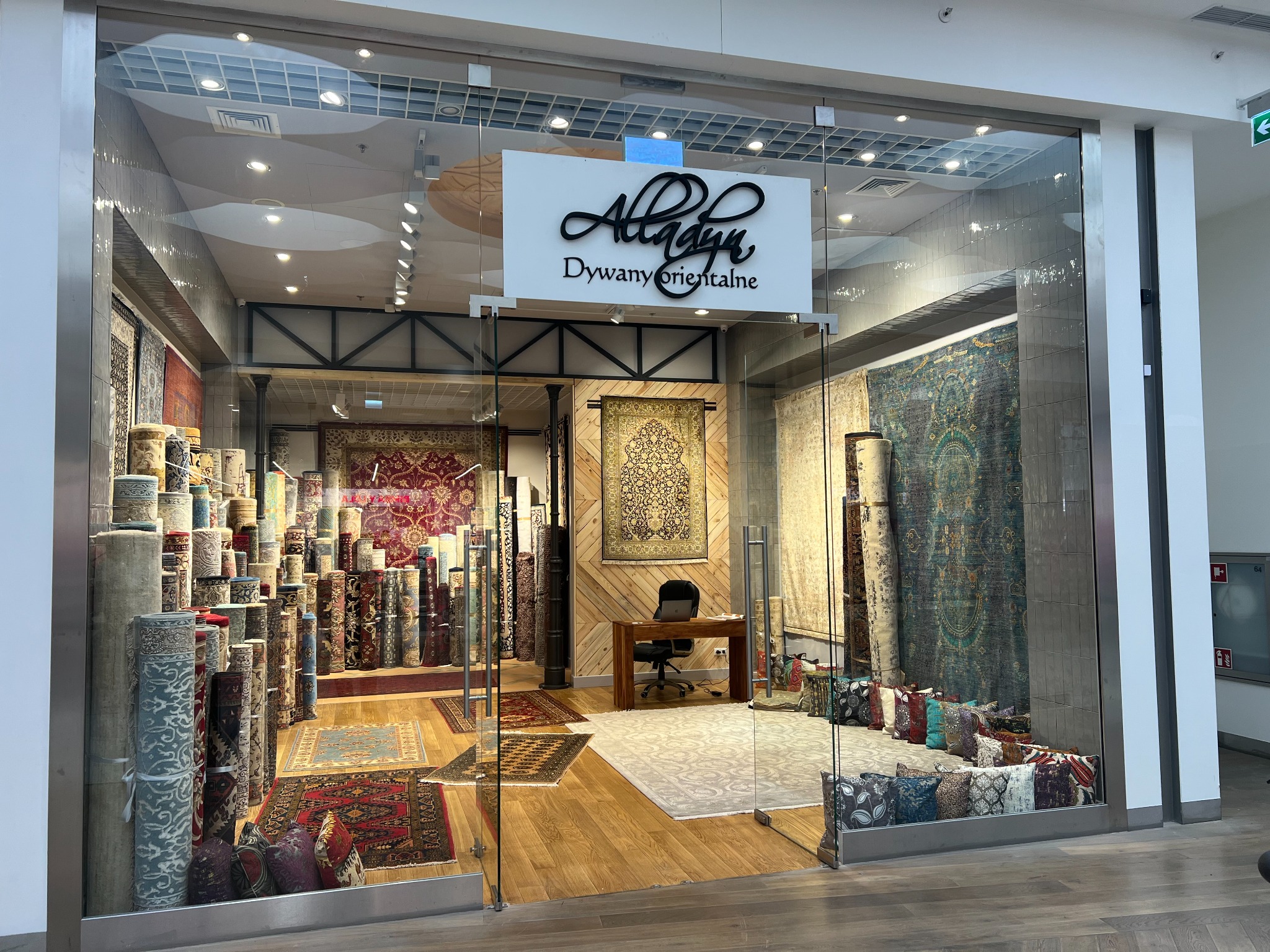 Wnętrze sklepu z dywanami orientalnymi za szklaną witryną, widoczne zwinięte dywany ustawione jeden na drugim, dywany rozłożone na podłodze i poduszki dekoracyjne przy ścianie, w głębi biurko...
