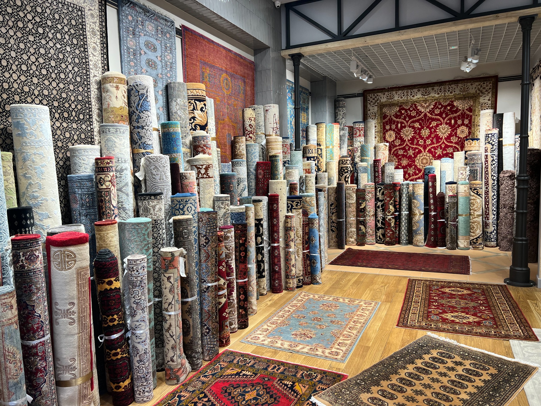 Wnętrze sklepu z dywanami: stosy zwiniętych wykładzin i dywanów z orientalnymi wzorami, ułożone na drewnianej podłodze, z dywanami rozwiniętymi na podłodze, oświetlenie punktowe.
