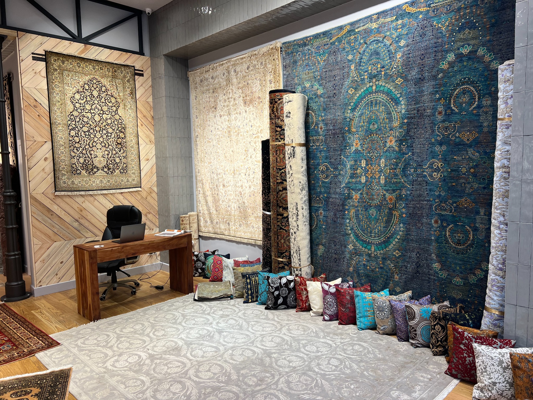 Salon z ekspozycją dywanów orientalnych, widoczne dywany wiszące na ścianach, zwinięte w rolki oraz rozłożone na podłodze, dodatkowo dekoracyjne poduszki i biurko z laptopem.