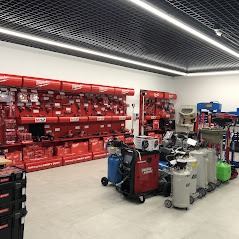 Wnętrze sklepu z narzędziami Milwaukee, ekspozycja na czerwonych półkach, widoczne kompresory i wózki narzędziowe na podłodze, oświetlenie sufitowe rastrowe.