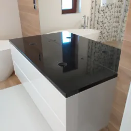 Czarny, połyskujący blat z otworami na baterię umywalkową, zamontowany na białej szafce łazienkowej z szufladami, w tle wanna i wzorzysta zasłona prysznicowa.