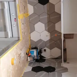 Układanie heksagonalnych płytek w odcieniach szarości i czerni na ścianie w kuchni, widoczne krzyżyki dystansowe, poziomica, wiadro z klejem i rolka papieru zabezpieczającego.