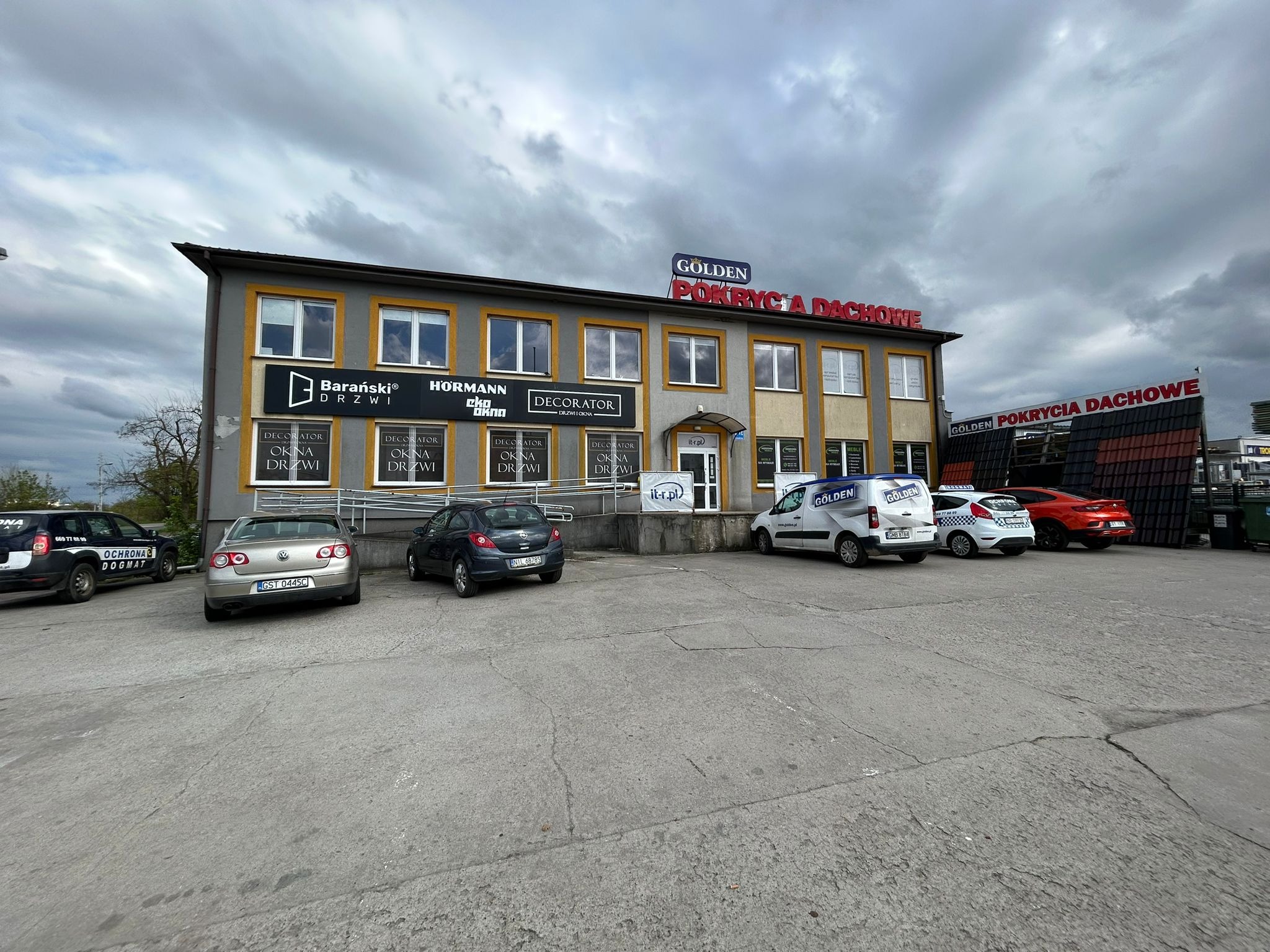 Dwupiętrowy budynek firmy Golden Pokrycia Dachowe z reklamami okien i drzwi, zaparkowane samochody firmowe na parkingu przed budynkiem w pochmurny dzień.