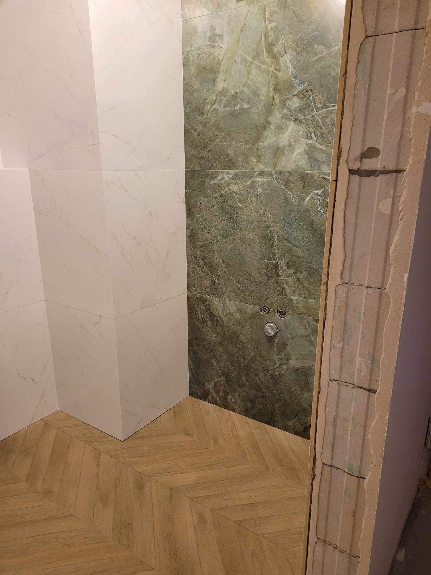 Fragment łazienki w trakcie remontu w Warszawie: ułożona podłoga z paneli imitujących drewno w jodełkę, częściowo wykończone ściany z białych i zielonych płytek imitujących marmur, widoczne surowe...