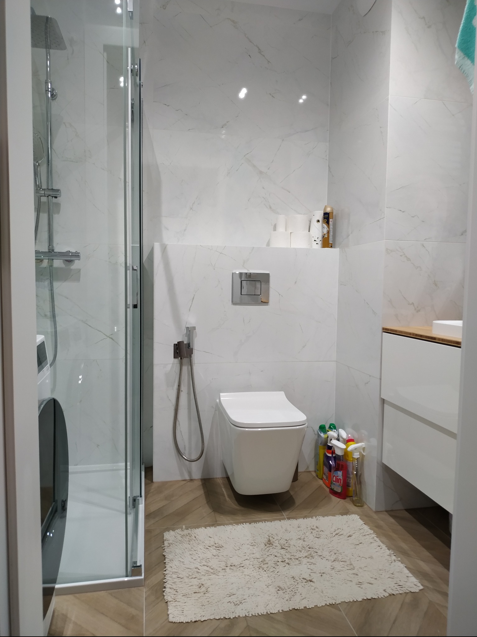 Nowoczesna łazienka z kabiną prysznicową typu walk-in, podwieszaną toaletą, marmurowymi płytkami ściennymi i drewnopodobnymi płytkami podłogowymi ułożonymi w jodełkę. Na podłodze leży puszysty...