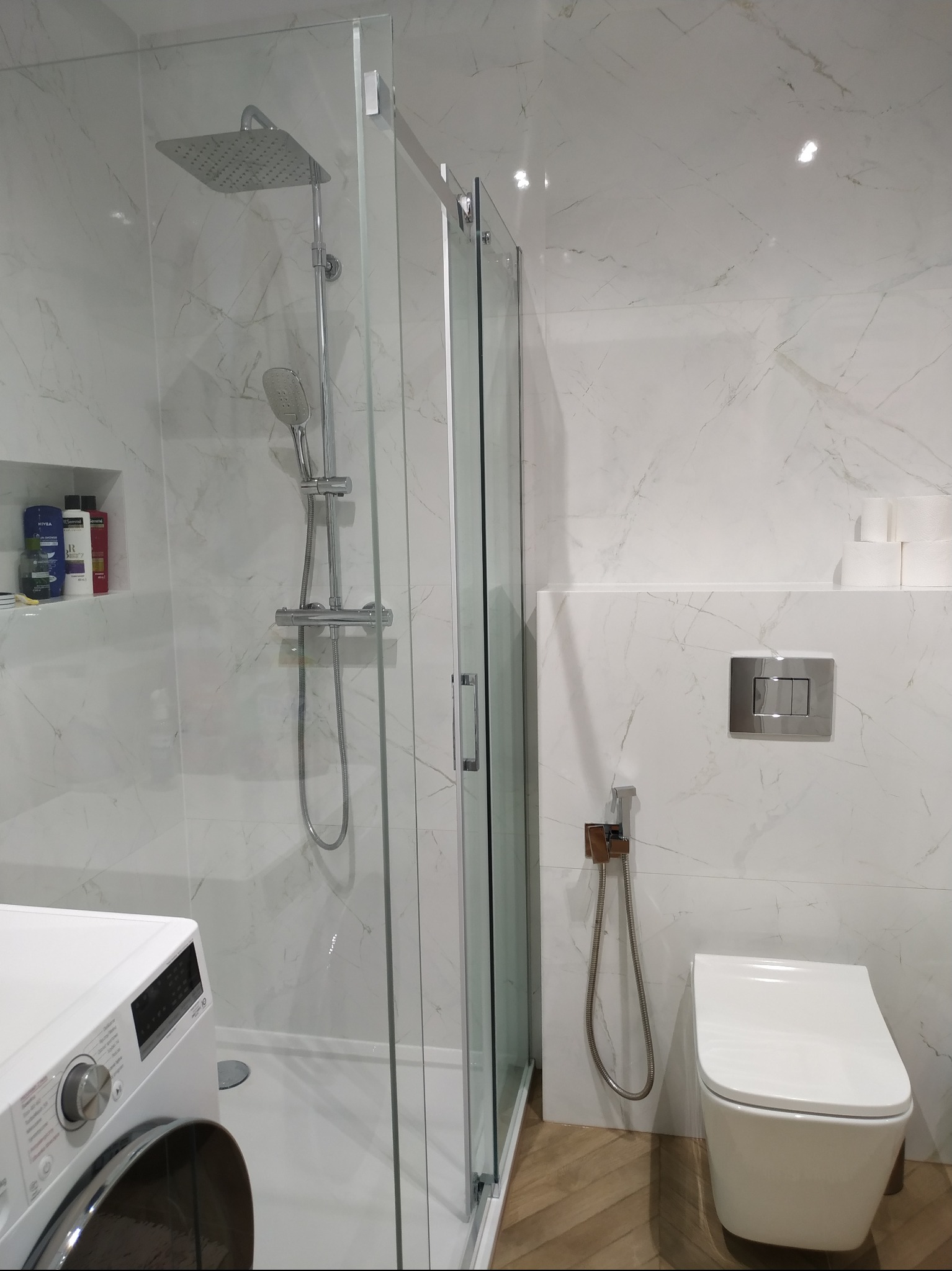 Nowoczesna łazienka z kabiną prysznicową typu walk-in, białe marmurowe płytki na ścianach, toaleta podwieszana z bidetką, pralka i półka z kosmetykami.
