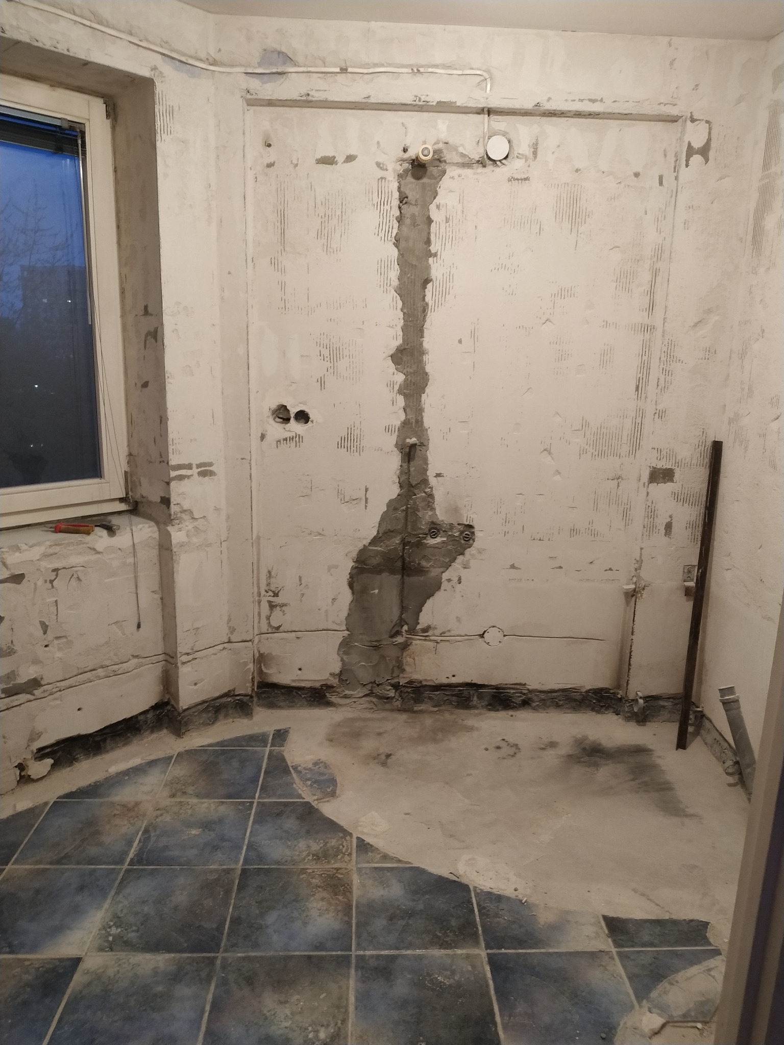 Stan surowy łazienki w trakcie prac: zerwane płytki, ściany z widocznymi śladami po skuciu, odkryte przewody elektryczne i rury, częściowo zachowana stara, ciemna glazura na podłodze.