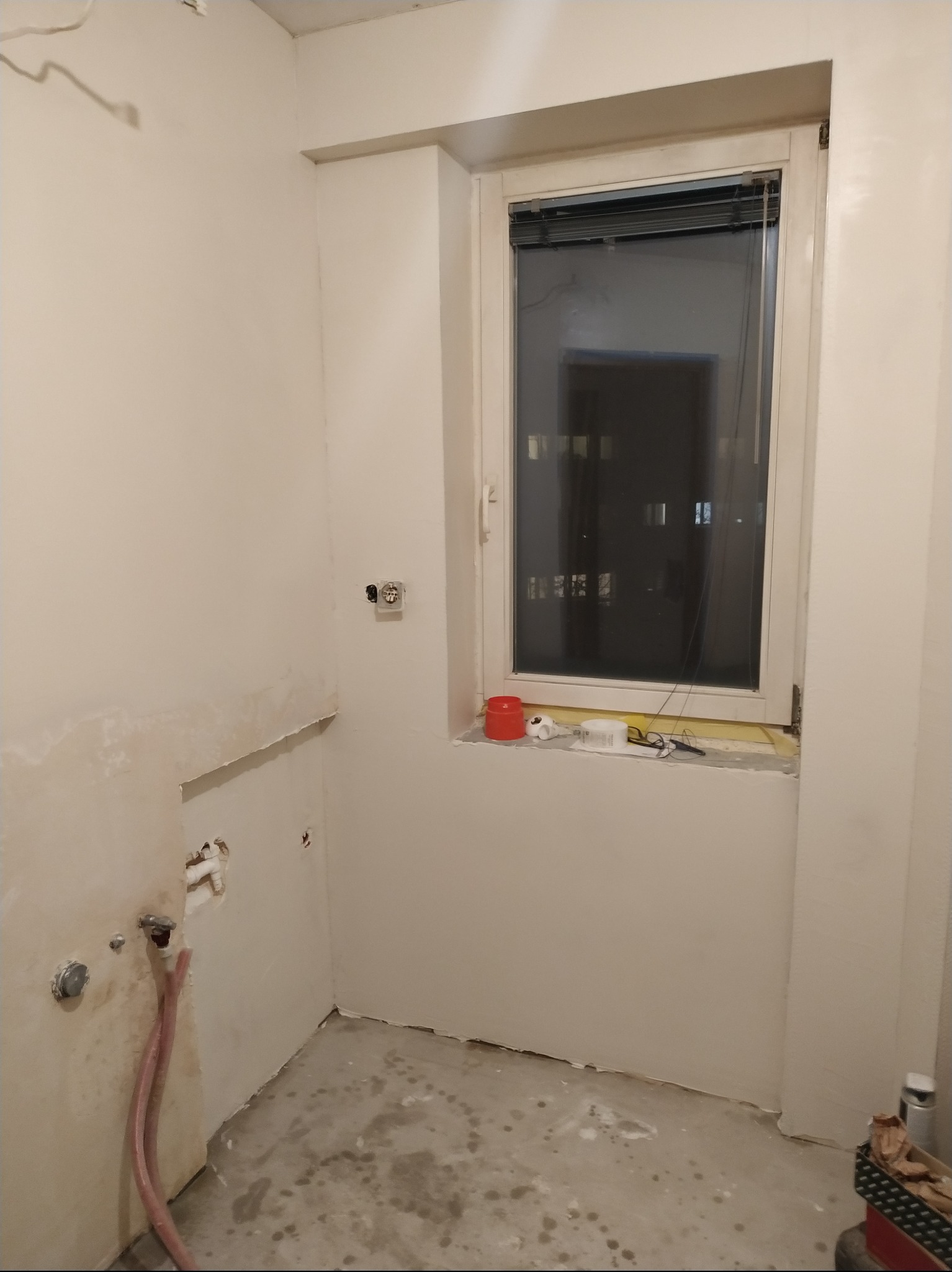 Stan surowy łazienki w trakcie przygotowań do remontu: ściany częściowo przygotowane, widoczne instalacje wodne, okno z roletami i narzędziami na parapecie.