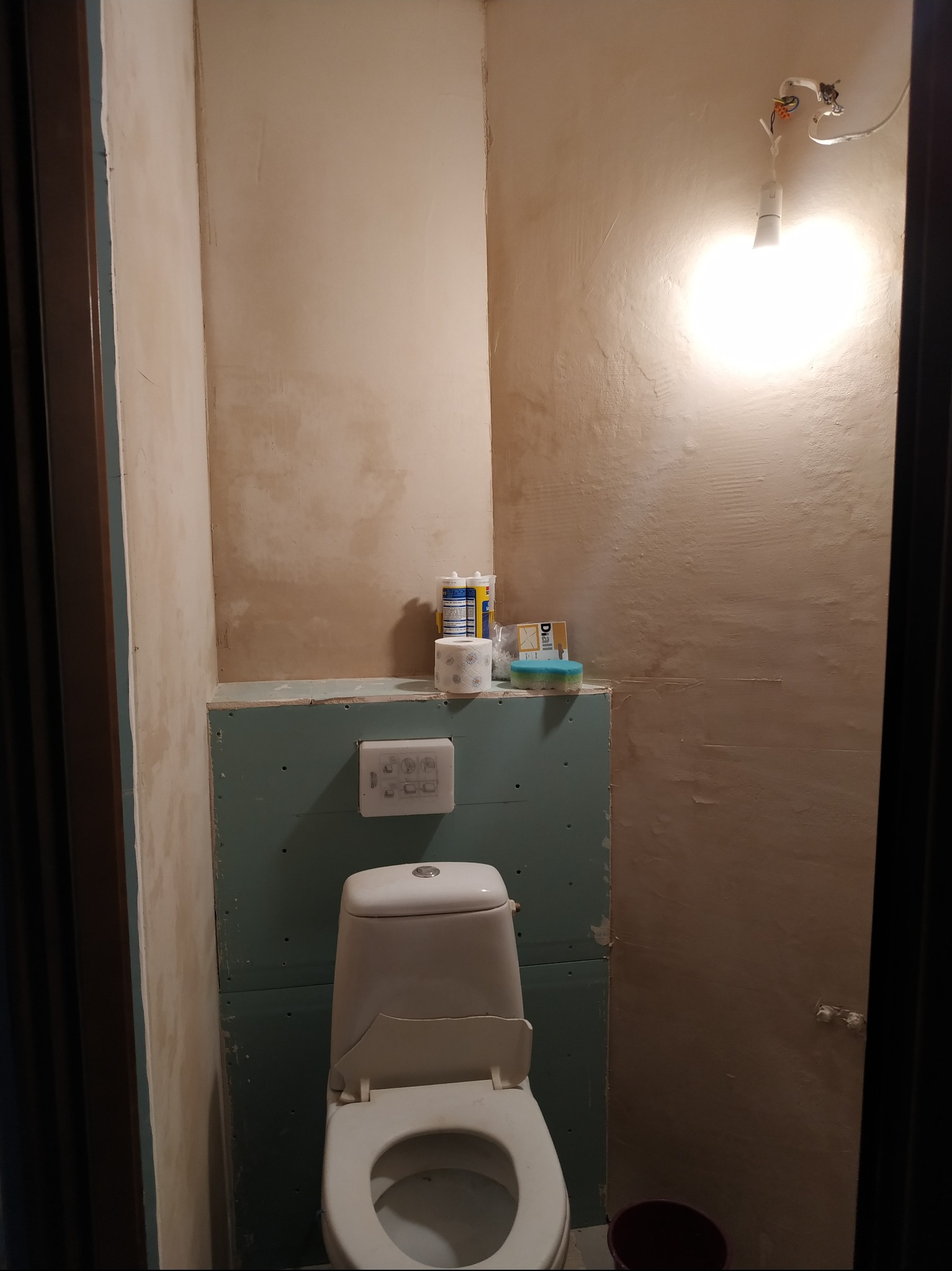 Niedokończona łazienka z toaletą, ściany w surowym tynku, widoczne płyty gipsowo-kartonowe, prowizoryczne oświetlenie żarówką