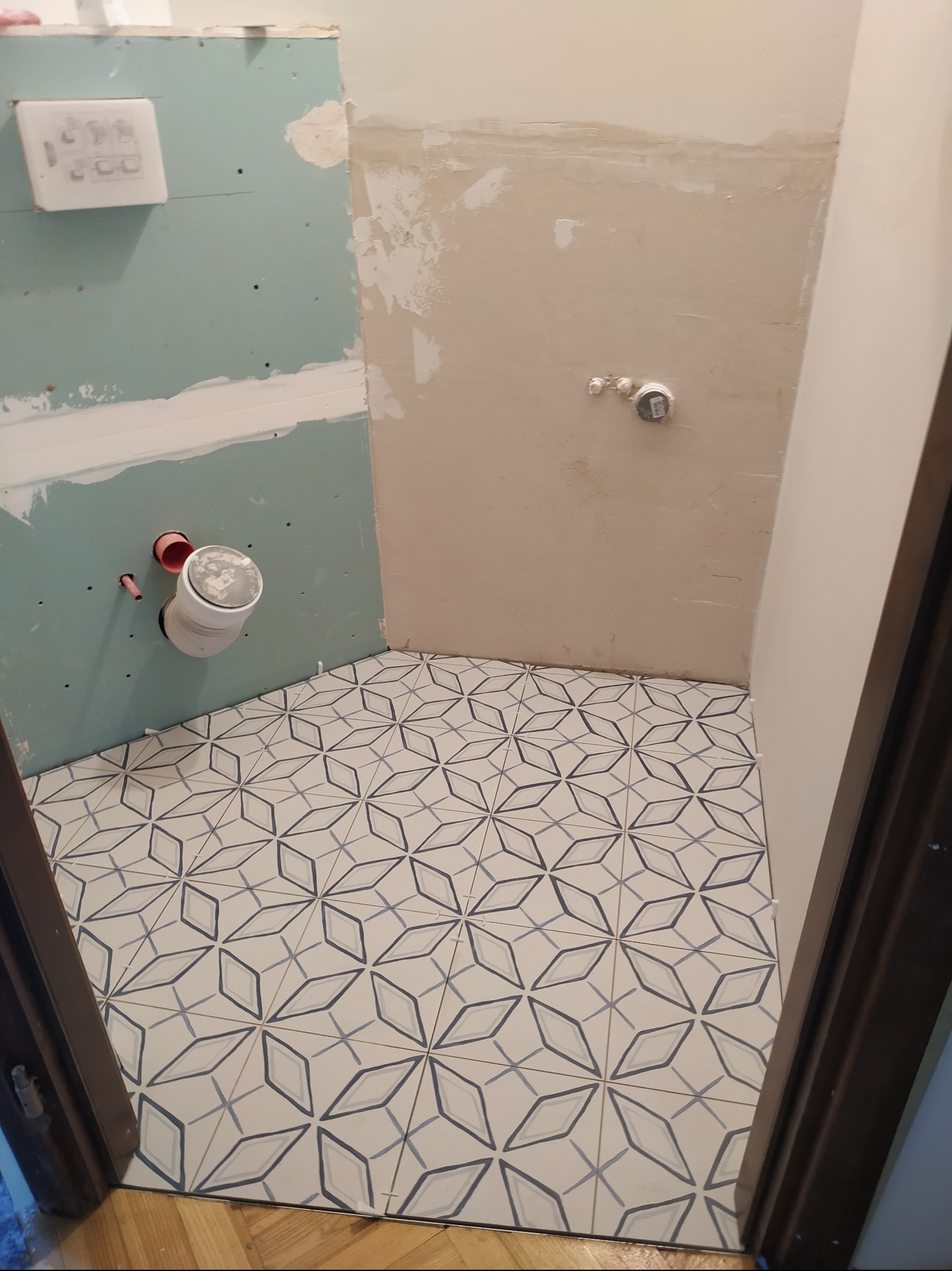 Fragment łazienki w trakcie prac: ułożona podłoga z geometrycznym wzorem, ściany częściowo wykończone płytami gipsowo-kartonowymi i gładzią, widoczne instalacje hydrauliczne.