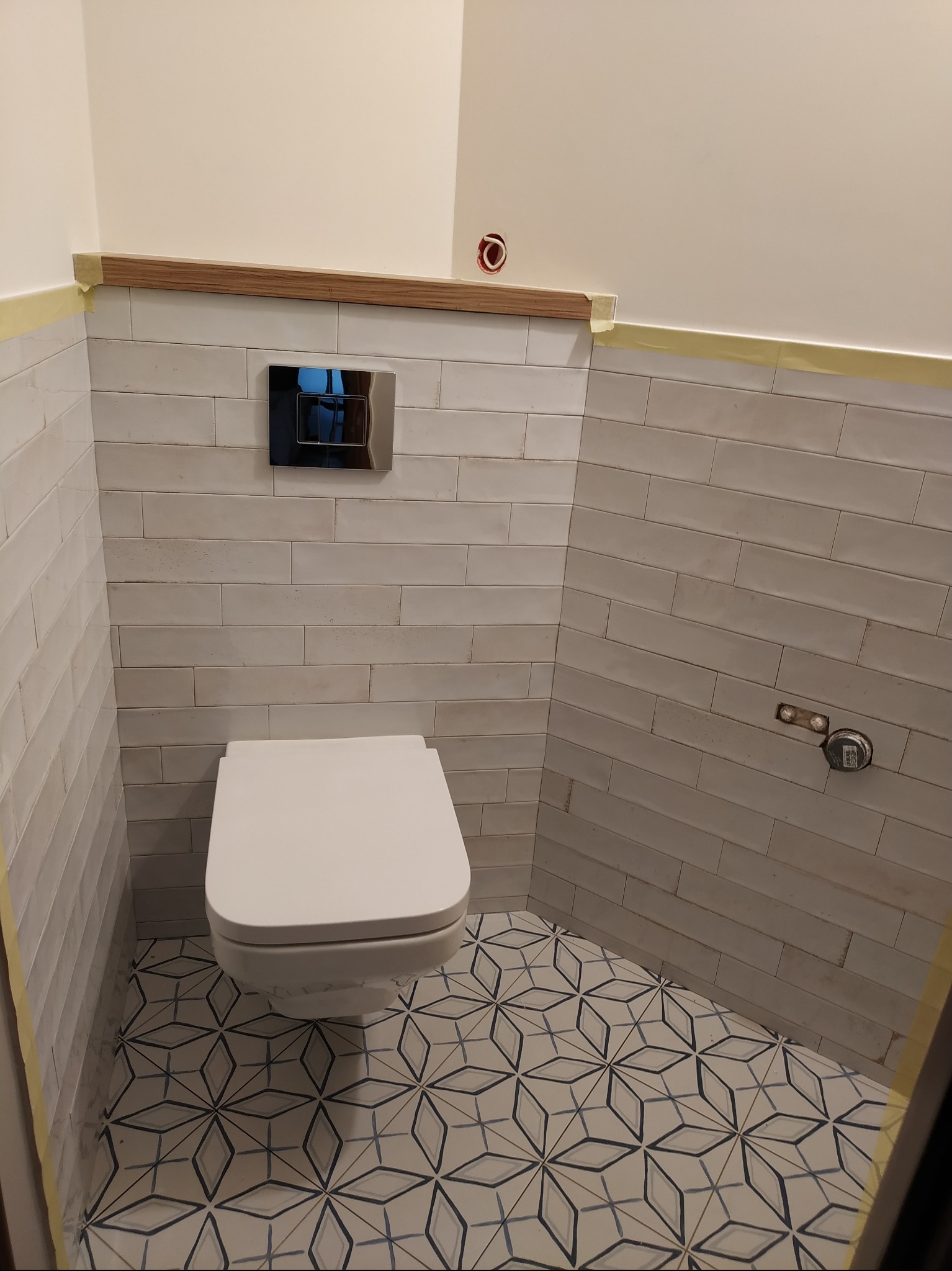 Nowoczesna toaleta wisząca w łazience z białymi, podłużnymi płytkami na ścianach i wzorzystą podłogą; widoczne elementy instalacji i taśma malarska zabezpieczająca krawędzie.