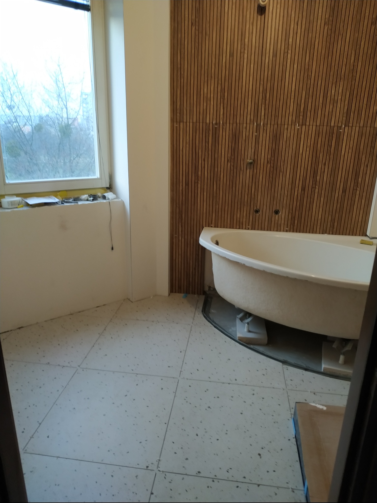 Łazienka w trakcie remontu: wanna na podkładkach, drewniana okładzina ścienna, okno z widokiem na drzewa, biała ściana z narzędziami na parapecie