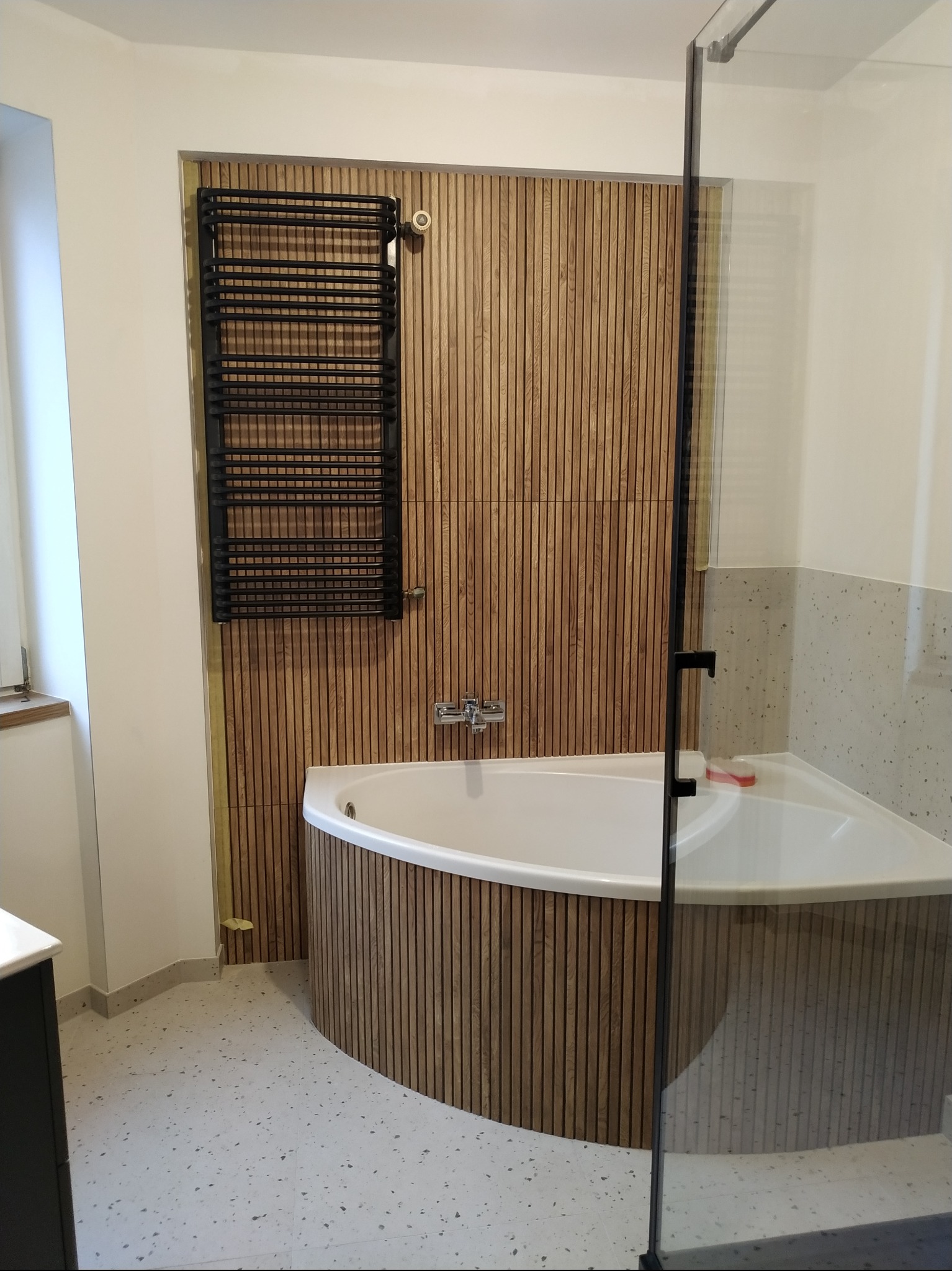 Nowoczesna łazienka z wanną narożną obudowaną drewnianymi lamelami, czarny grzejnik drabinkowy na ścianie z lamelami, szklana kabina prysznicowa z czarnymi okuciami, podłoga w terazzo.