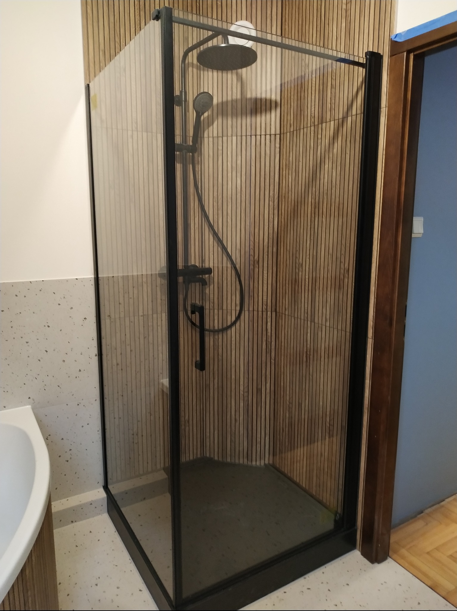 Nowoczesna kabina prysznicowa z czarnymi profilami i deszczownicą na tle ściany wyłożonej drewnianymi panelami w łazience z podłogą w kolorze terazzo.