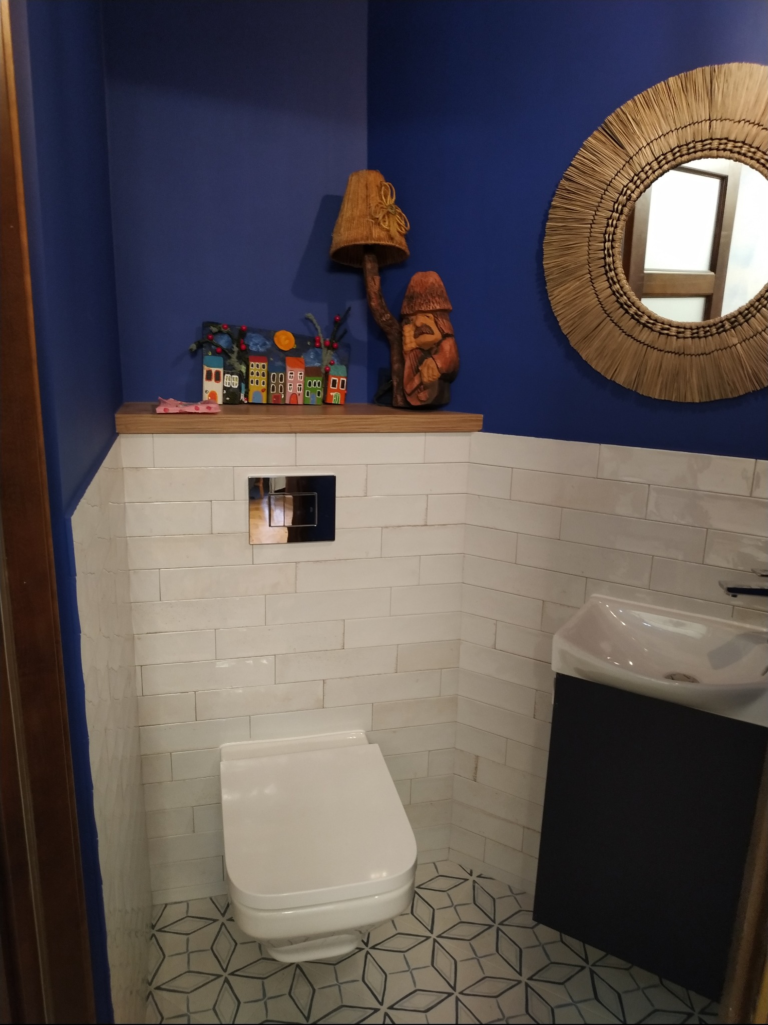 Niebieska ściana łazienki z białymi, prostokątnymi płytkami, toaleta z podwieszanym systemem spłukiwania Geberit, okrągłe lustro z rafii, dekoracje na półce i wzorzysta podłoga.