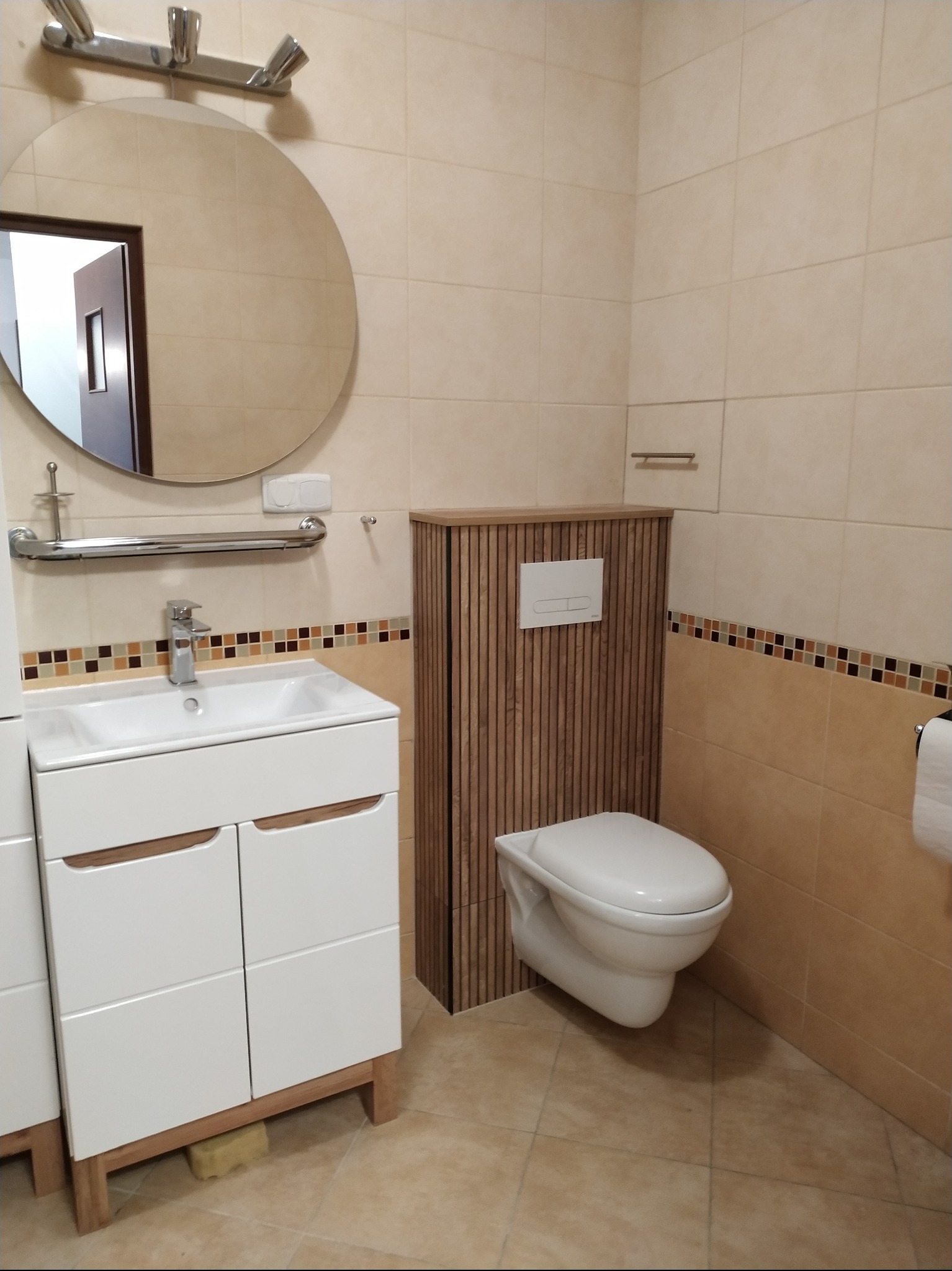 Jasna łazienka z beżowymi płytkami, białą umywalką z szafką, okrągłym lustrem i podwieszanym WC z zabudową z drewnianych listewek.