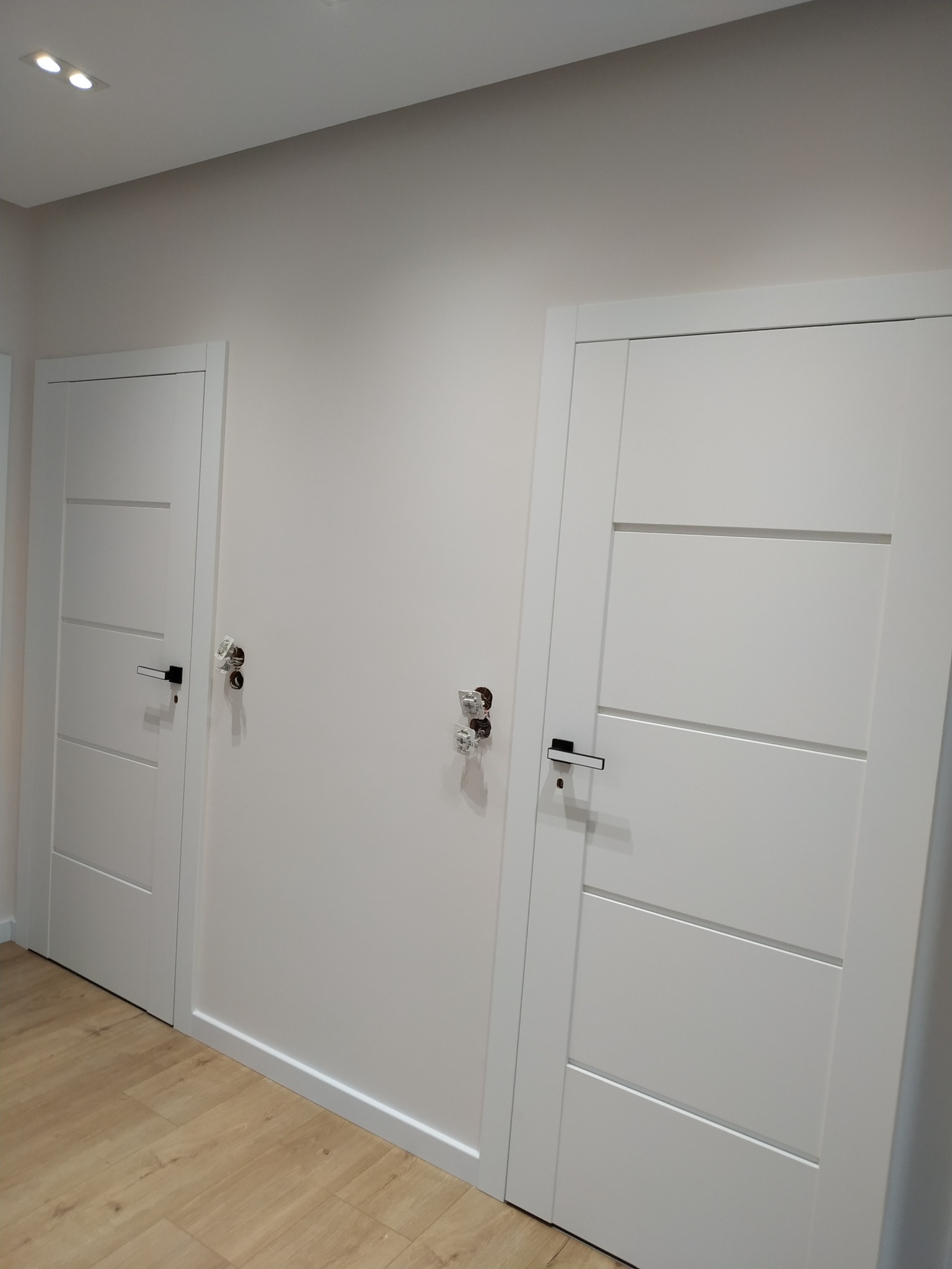 Dwa białe, nowoczesne drzwi wewnętrzne z czarnymi klamkami, zamontowane w jasnym pomieszczeniu z drewnopodobną podłogą; widoczne otwory po poprzednich klamkach.