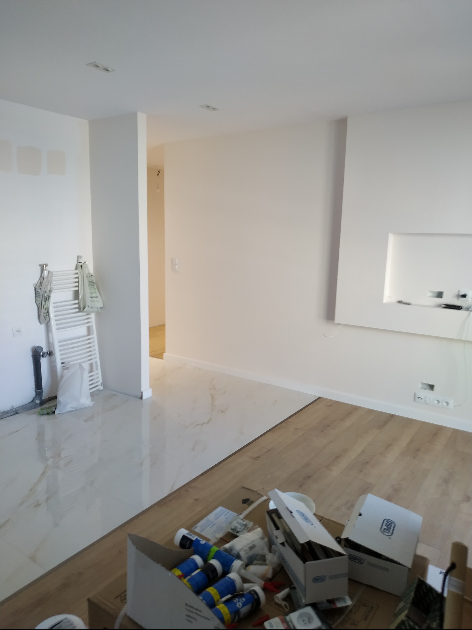 Wnętrze w trakcie prac wykończeniowych: połączenie płytek imitujących marmur i paneli podłogowych, widoczne narzędzia i materiały budowlane w kartonach.