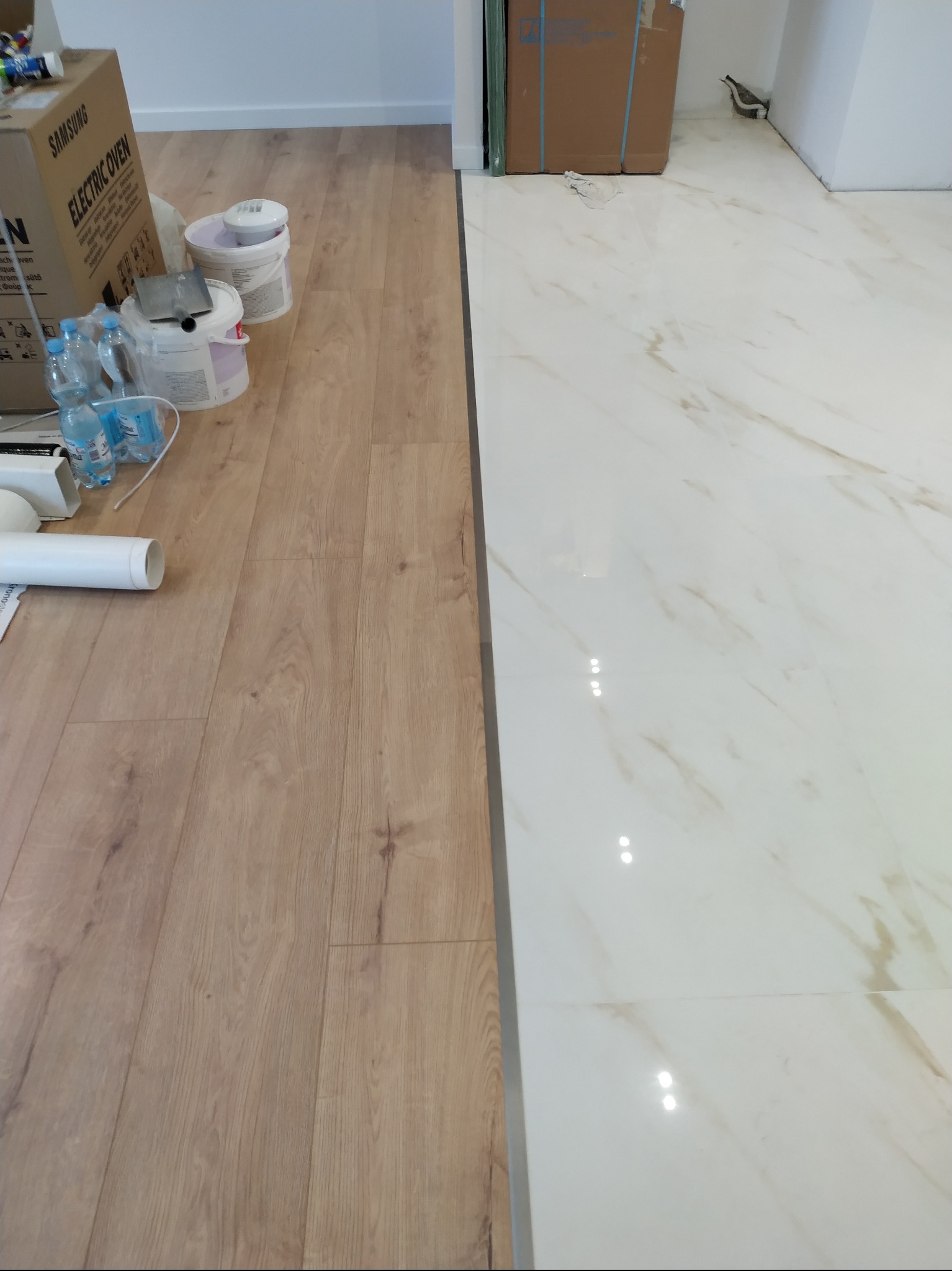 Połączenie jasnych paneli podłogowych z białymi płytkami imitującymi marmur, widoczne puszki z farbą i narzędzia, kartonowe pudło po piekarniku Samsung, butelki z wodą i rura PCV w trakcie prac...