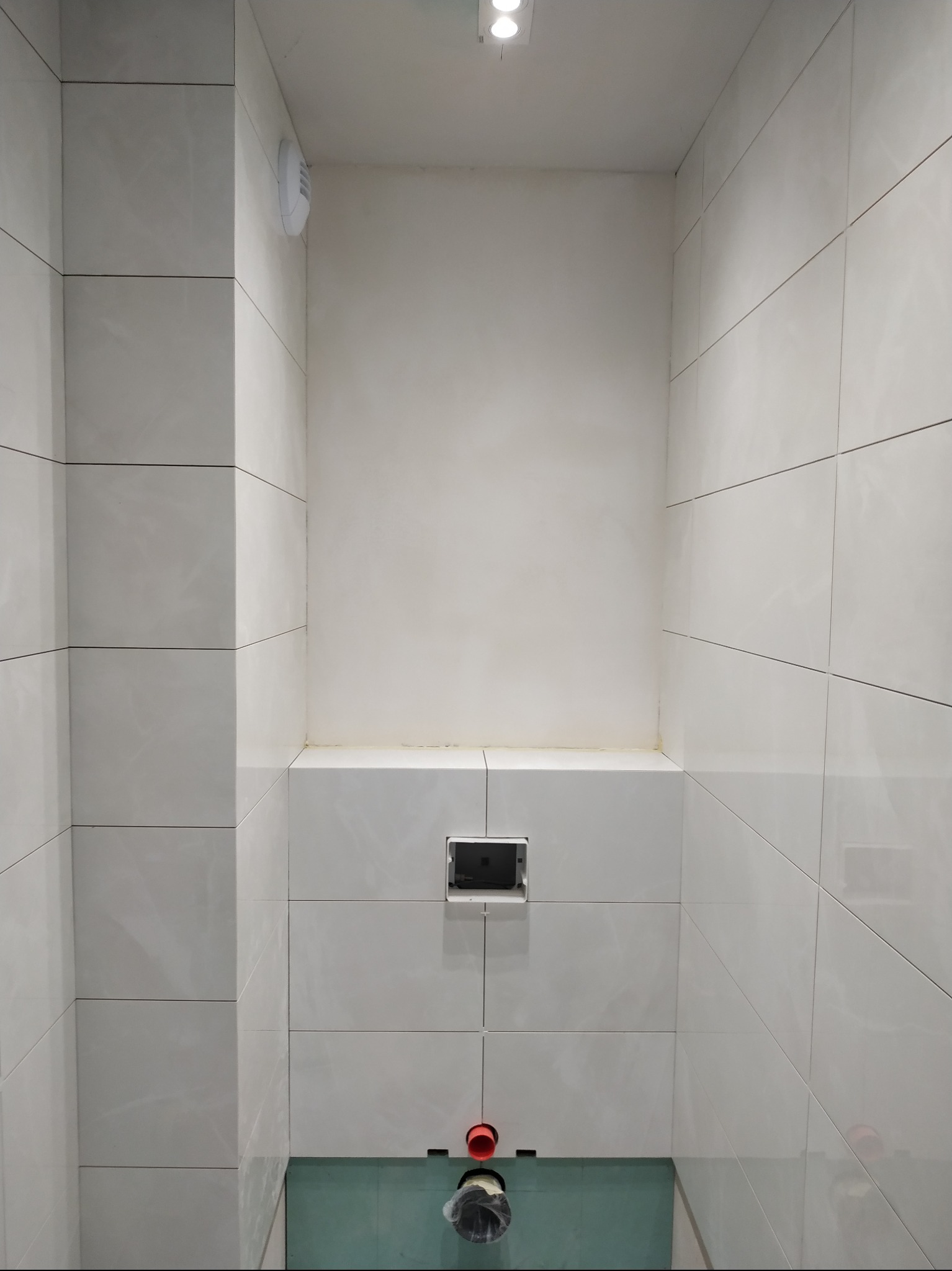 Wnętrze łazienki w trakcie prac wykończeniowych, widoczne białe płytki na ścianach, instalacja podtynkowa WC oraz fragment podłogi z zieloną izolacją.