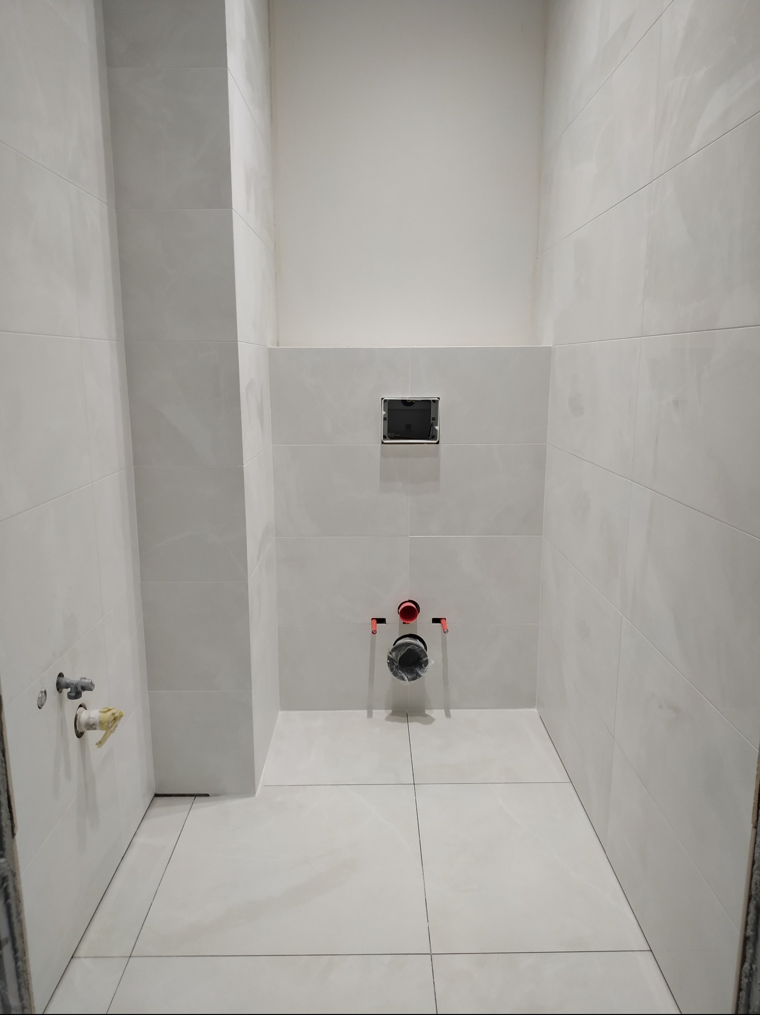 Wnętrze łazienki w trakcie prac wykończeniowych; widoczne białe płytki na ścianach i podłodze, zamontowany stelaż podtynkowy WC oraz przygotowane przyłącza wodno-kanalizacyjne.