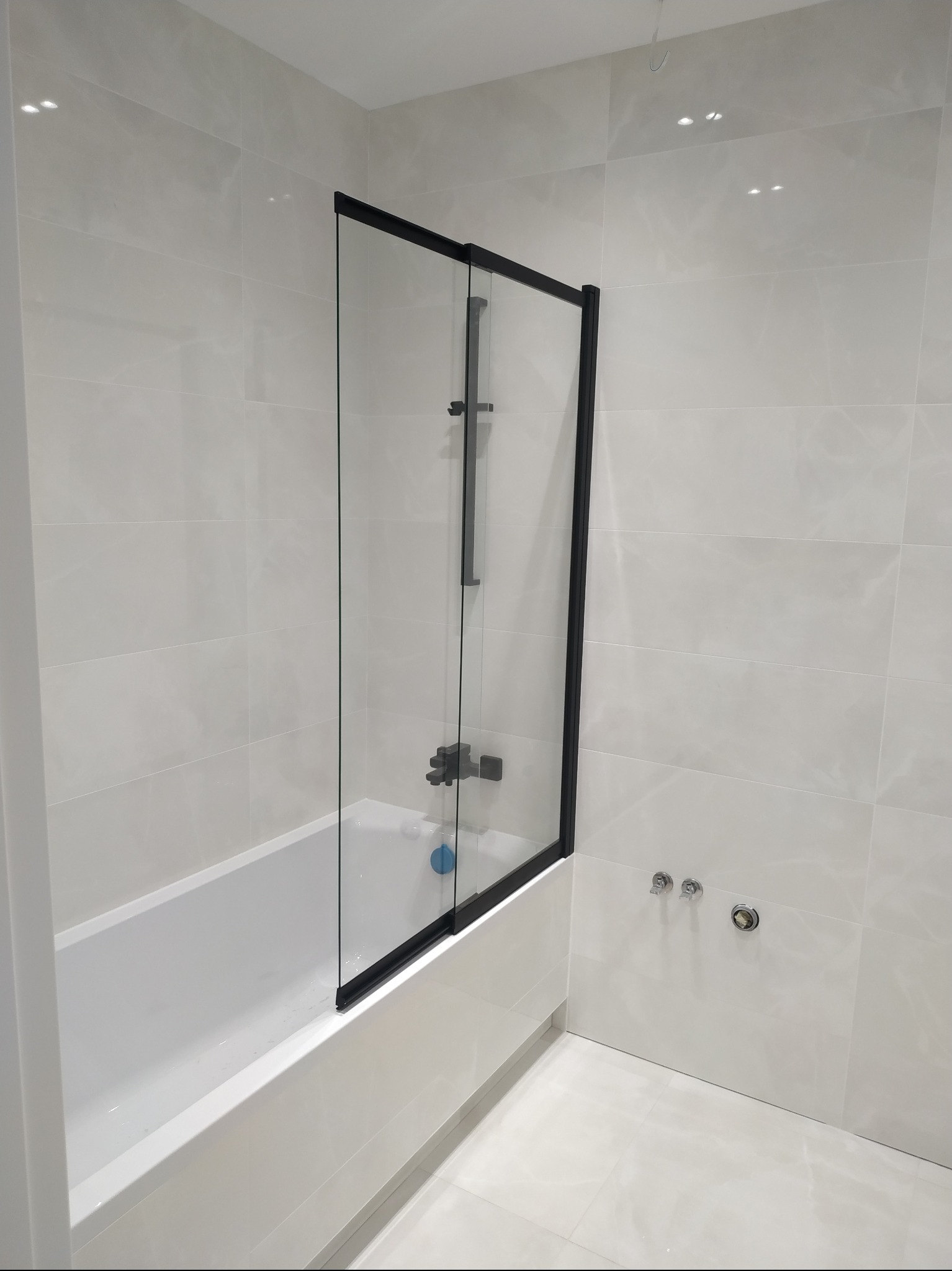 Nowoczesna łazienka z wanną i czarną, szklaną ścianką prysznicową, wykończona płytkami w odcieniach bieli i szarości, prezentująca minimalistyczny design.